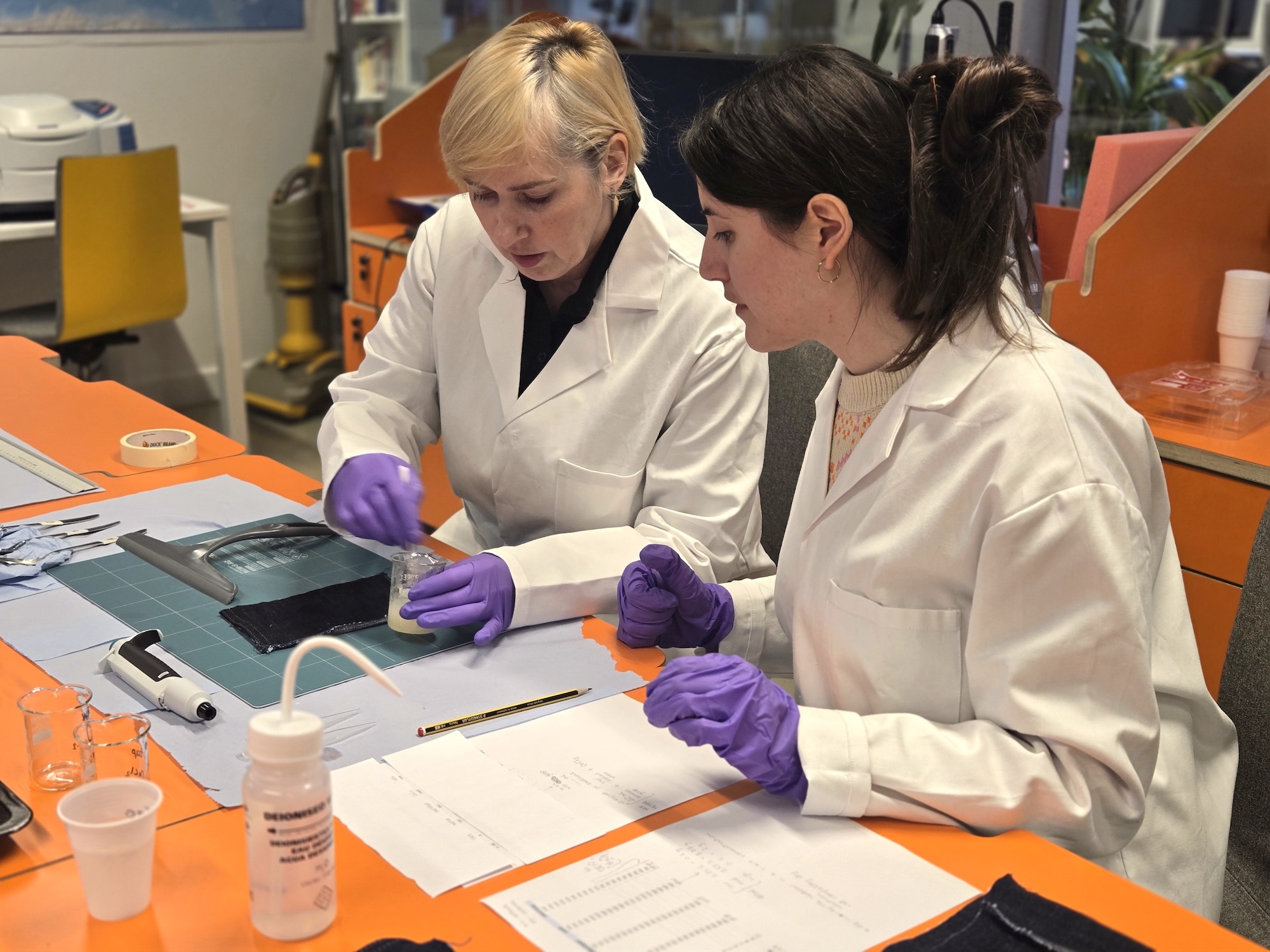 Two people in white lab coats and purple gloves work at an orange laboratory bench, examining samples and paperwork with glassware and a wash bottle nearby.