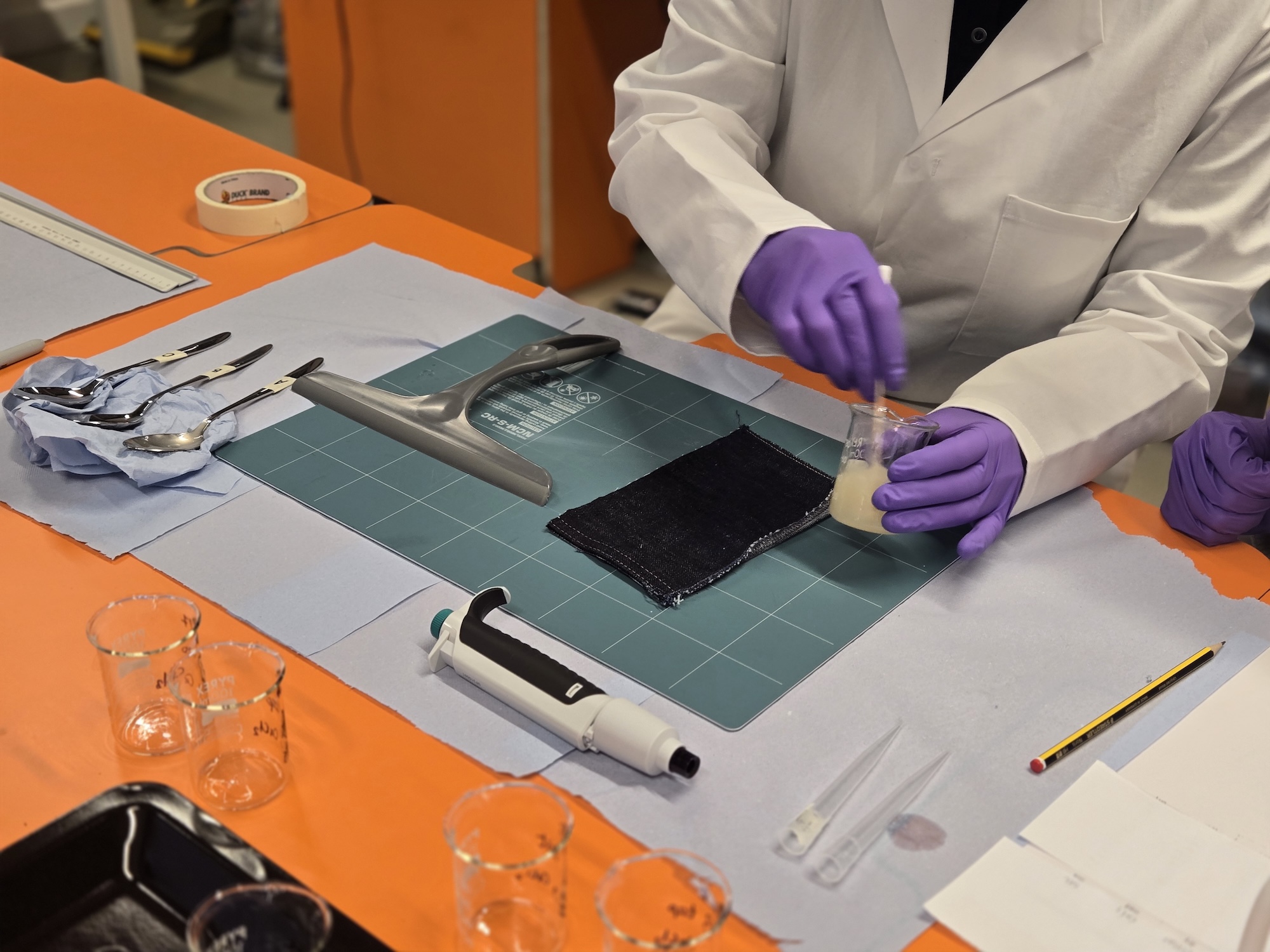 A lab worker in a white coat and purple gloves mixes a pale liquid in a small container on a cutting mat, with brushes, tape, a caliper and glass beakers arranged on an orange table.