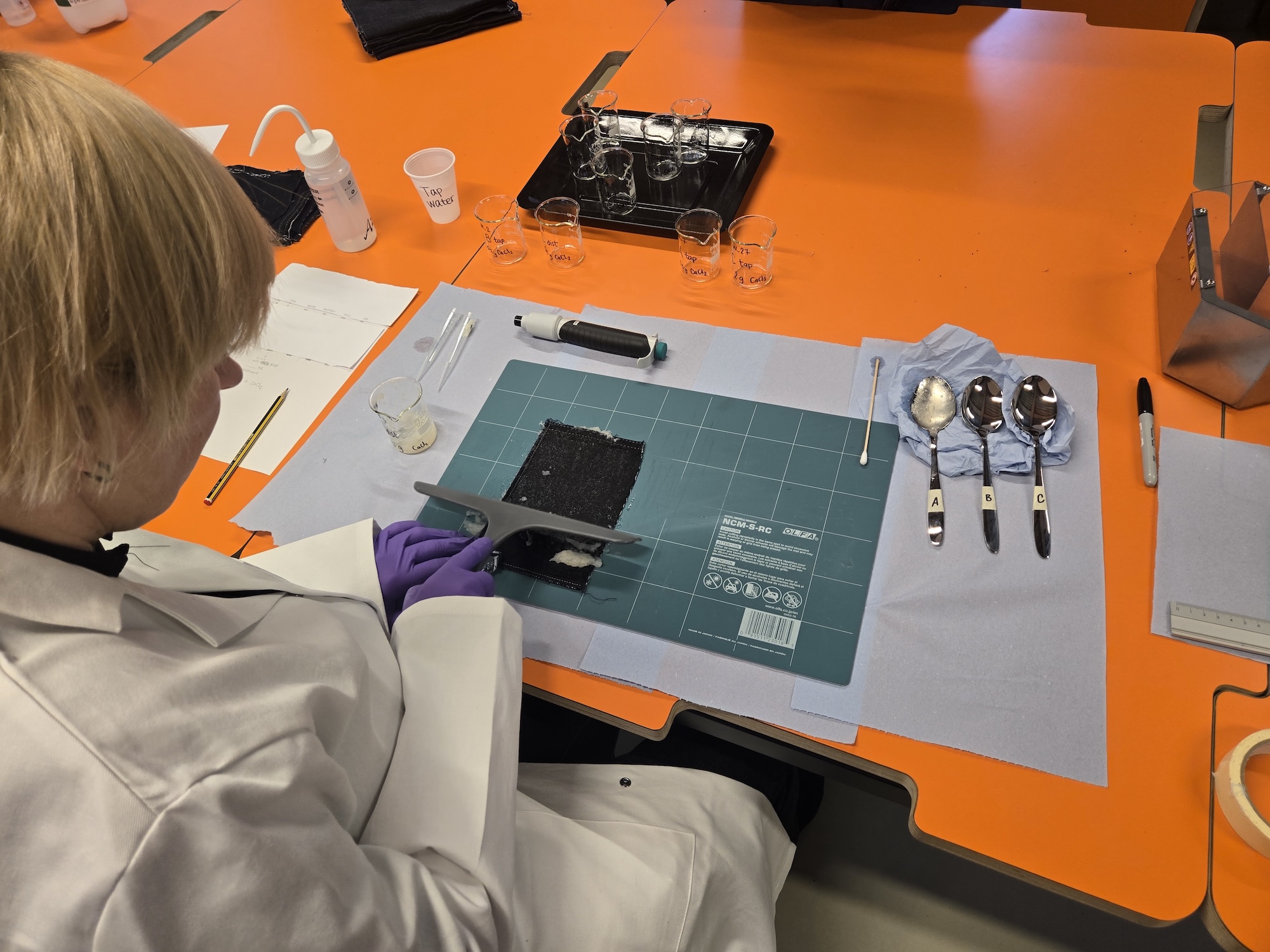 A person in a white lab coat and purple gloves working on a sample at a cutting mat, with spoons, glasses and lab tools on an orange table.