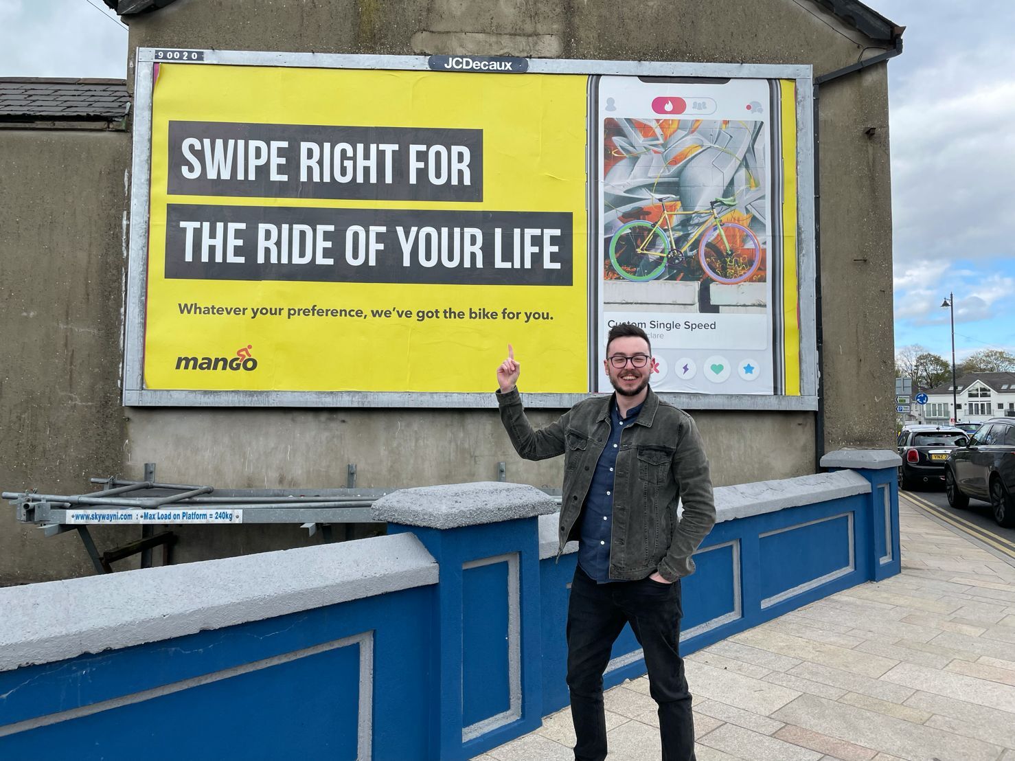 Man in glasses and green jacket pointing at yellow billboard advertising bikes, standing by blue railings on street