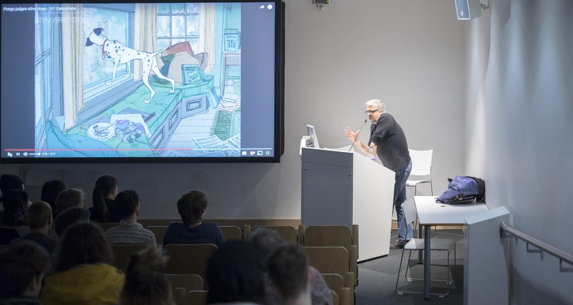 S Chomet delivers a lecture in a lecture theatre. A screen to the left shows part of a presentation.