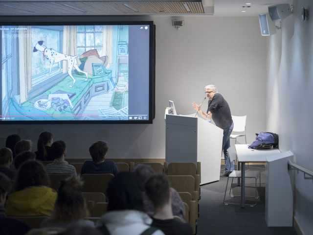 A man delivers a lecture in a lecture theatre. A screen to the left shows part of a presentation.