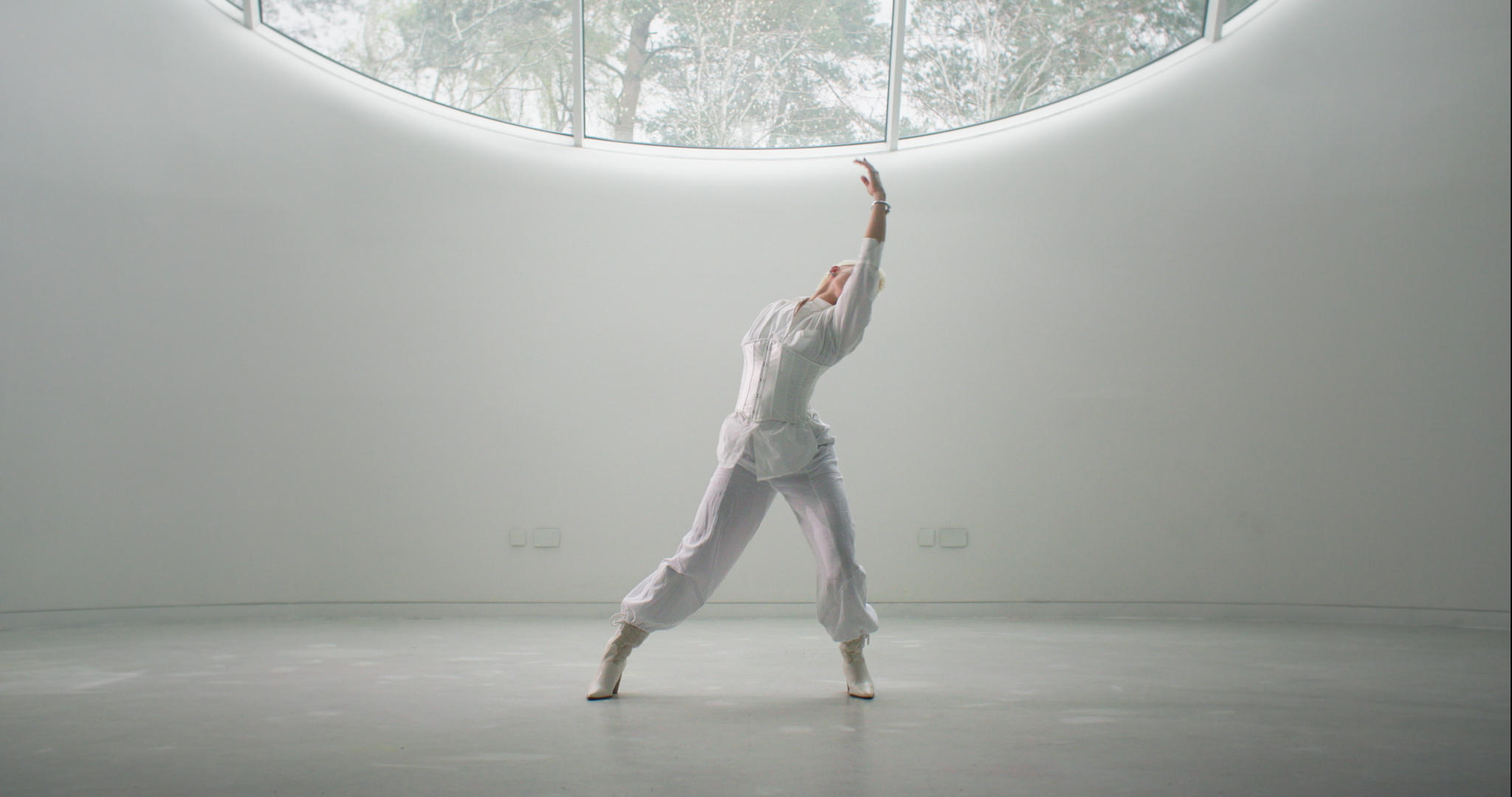 Ballet dancer in white outfit performing leap in curved white room with large window showing greenery outside.