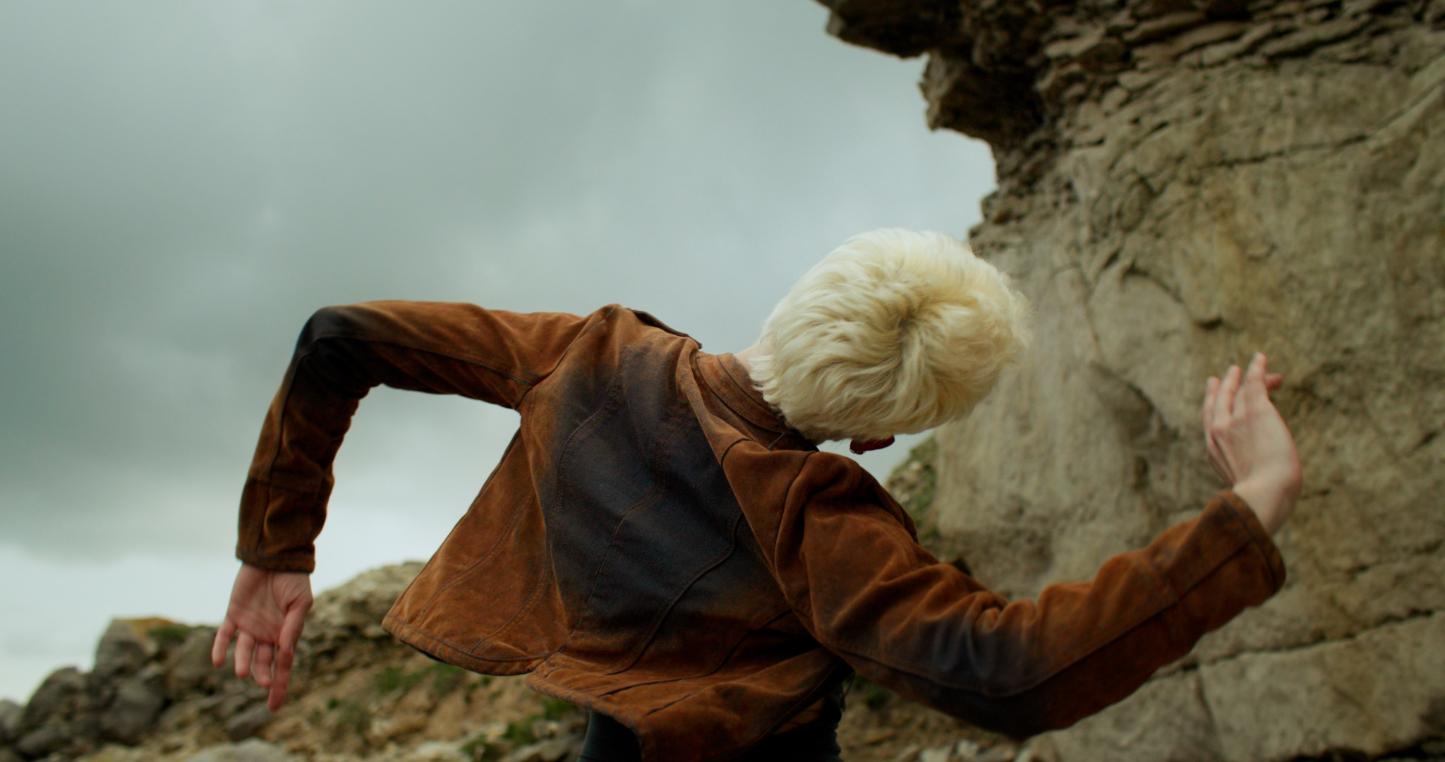 Person with blonde hair in orange jacket dancing in front of grey rock face, one hand in the air, overcast sky visible.