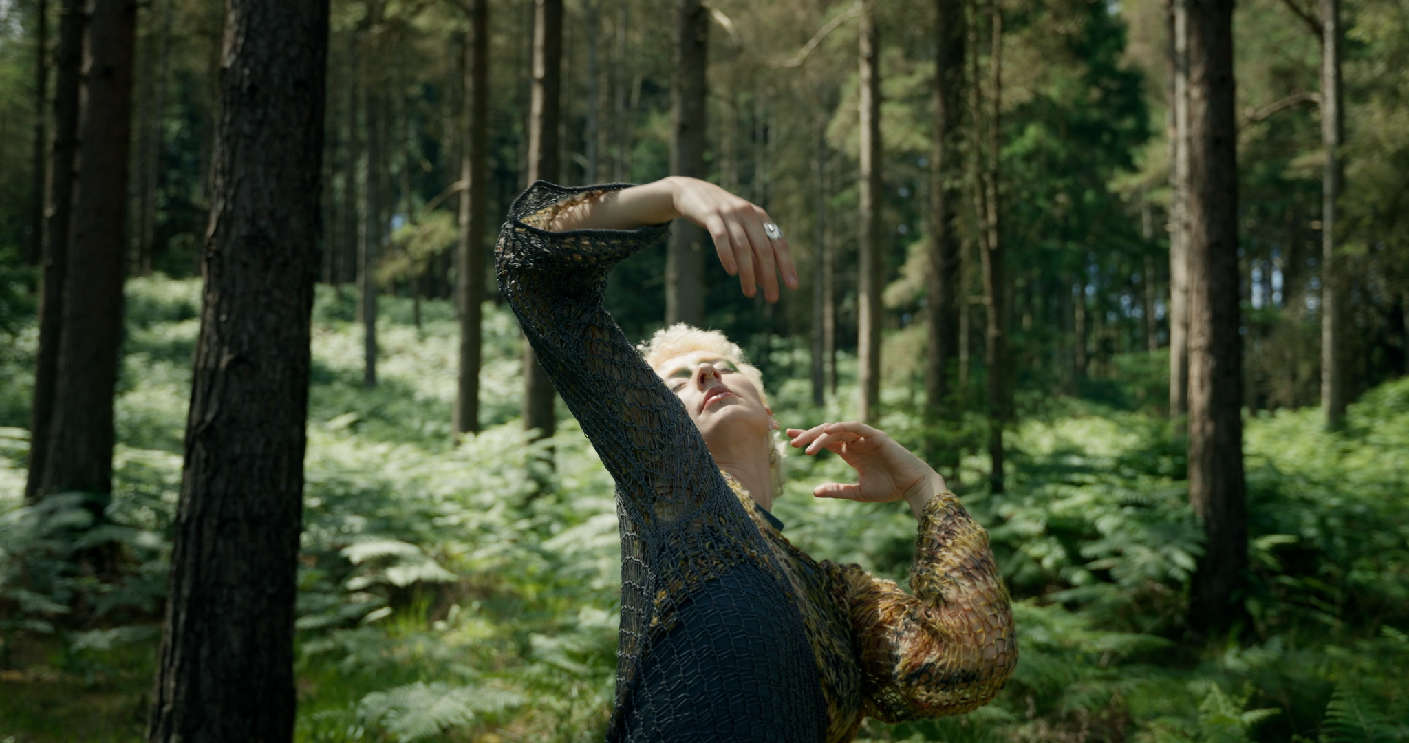 Dancer with green eyeshadow dark jumper raising one arm amongst tall forest trees with green undergrowth and dappled sunlight.