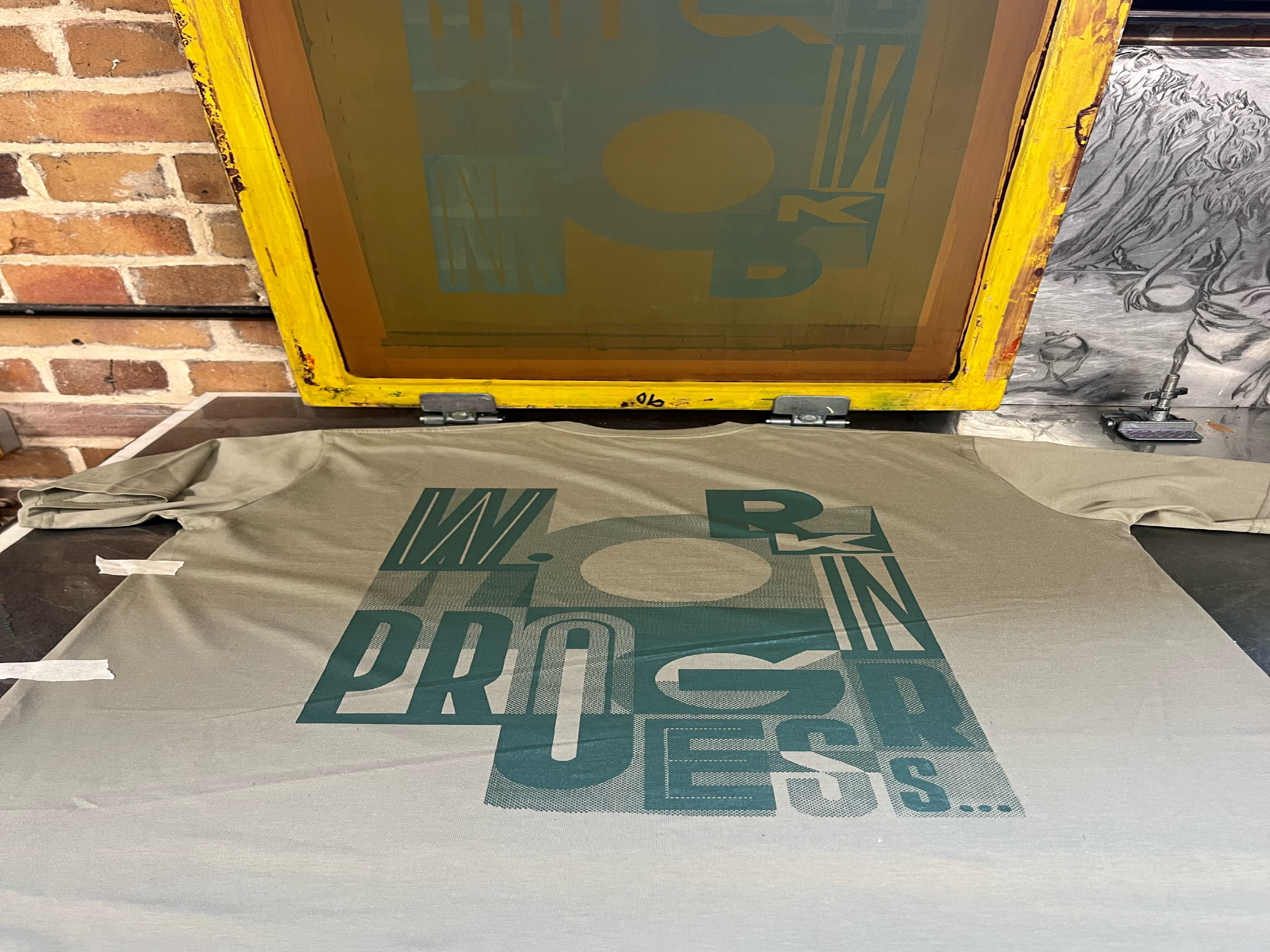 A white T-shirt with a screen-printed abstract lettering design. The design reads "Work in Progress" in mismatching fonts.