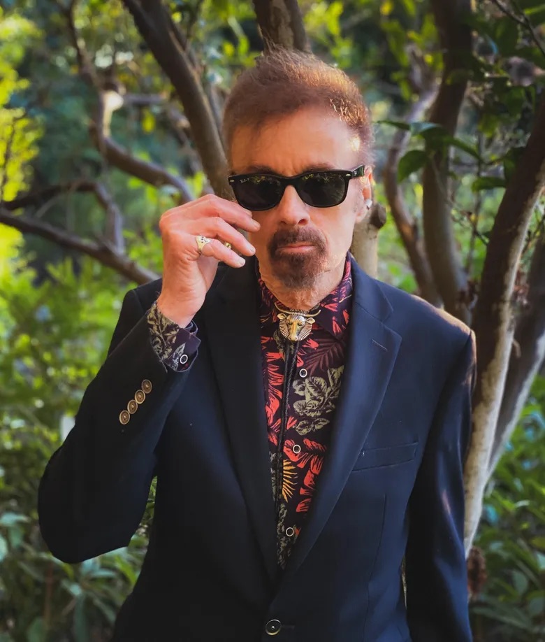 T.C. Boyle stood outside in forest, wearing sunglasses, floral shirt and dark blue blazer.