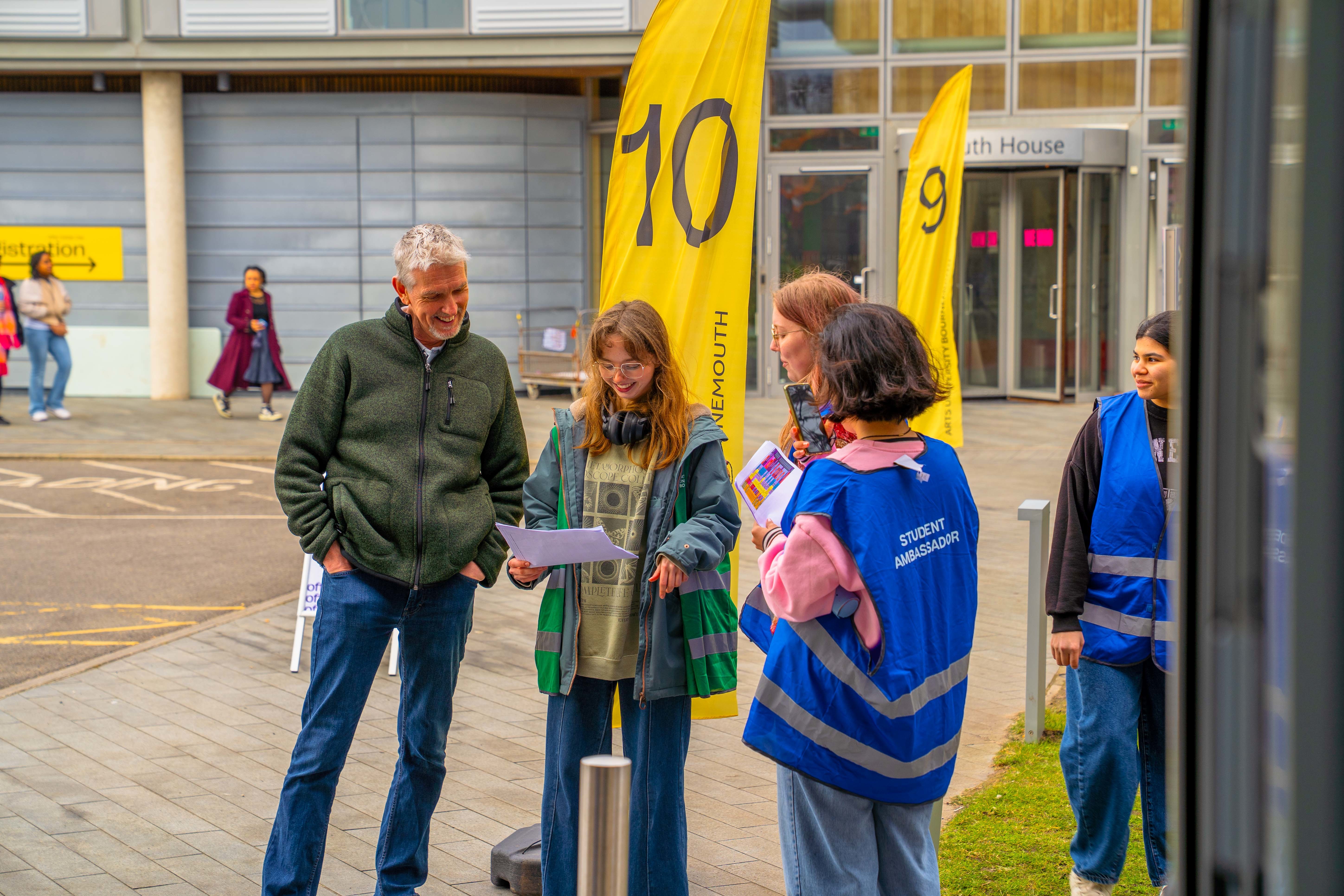 Two visitors to AUB stand in the courtyard on campus looking at a map while speaking to Student Ambassadors. Yellow numbered flags appear in the background.