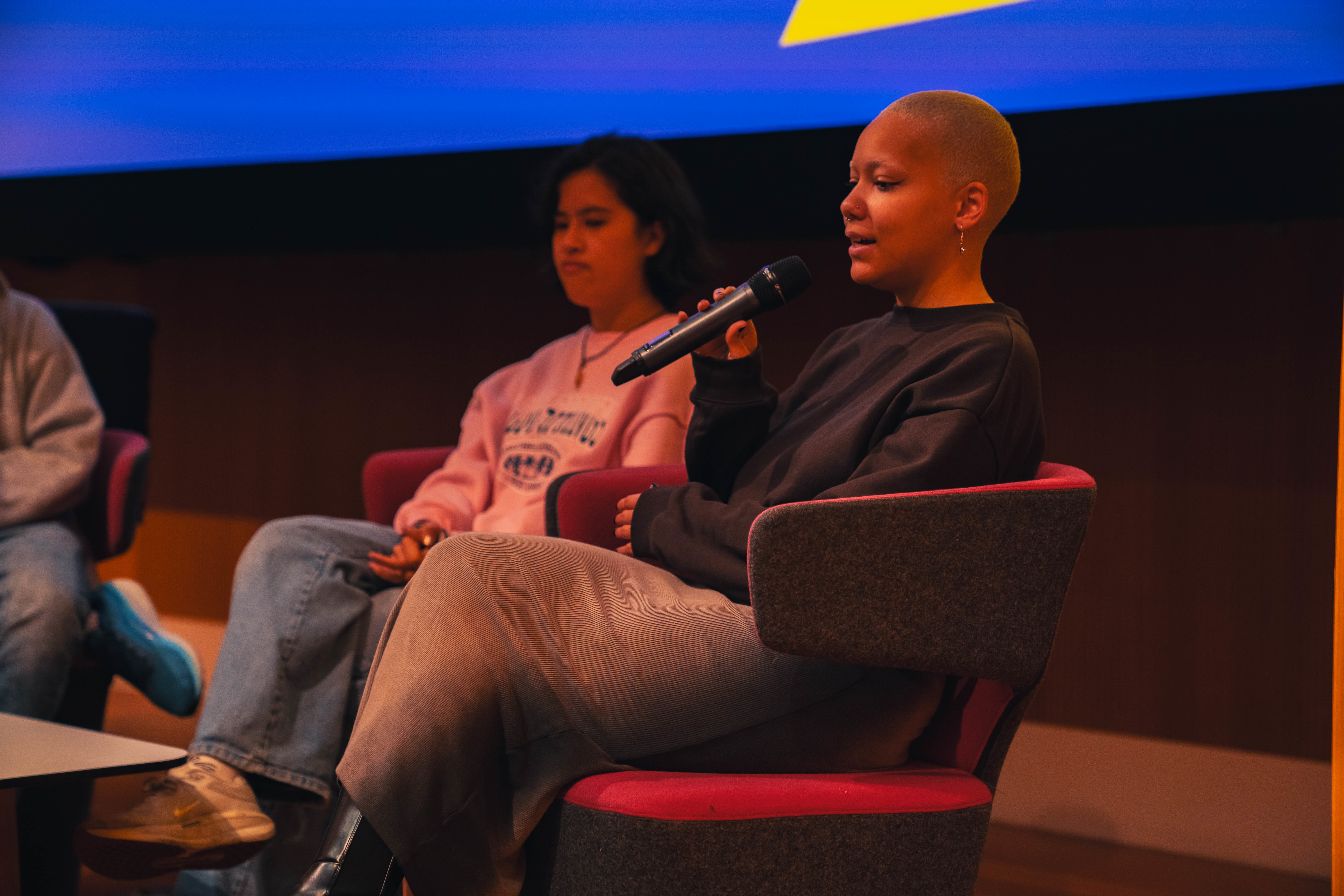 Two members of a student panel sitting in chairs on stage in an Arts University Bournemouth lecture theatre. One of the students, in the foreground, is speaking into a microphone.