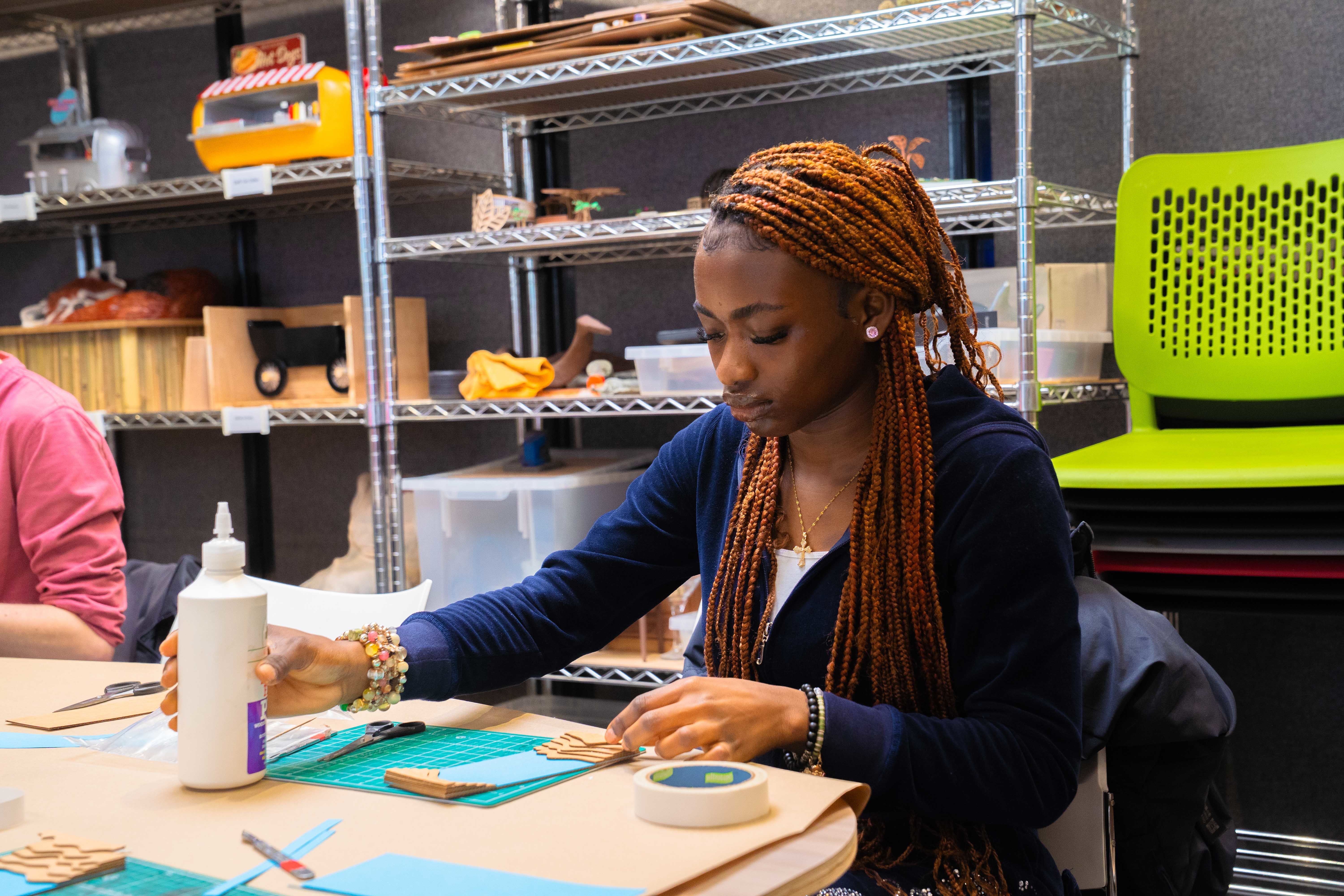 A young person sitting at a table in an AUB studio with art supplies and a cutting board in front of them.