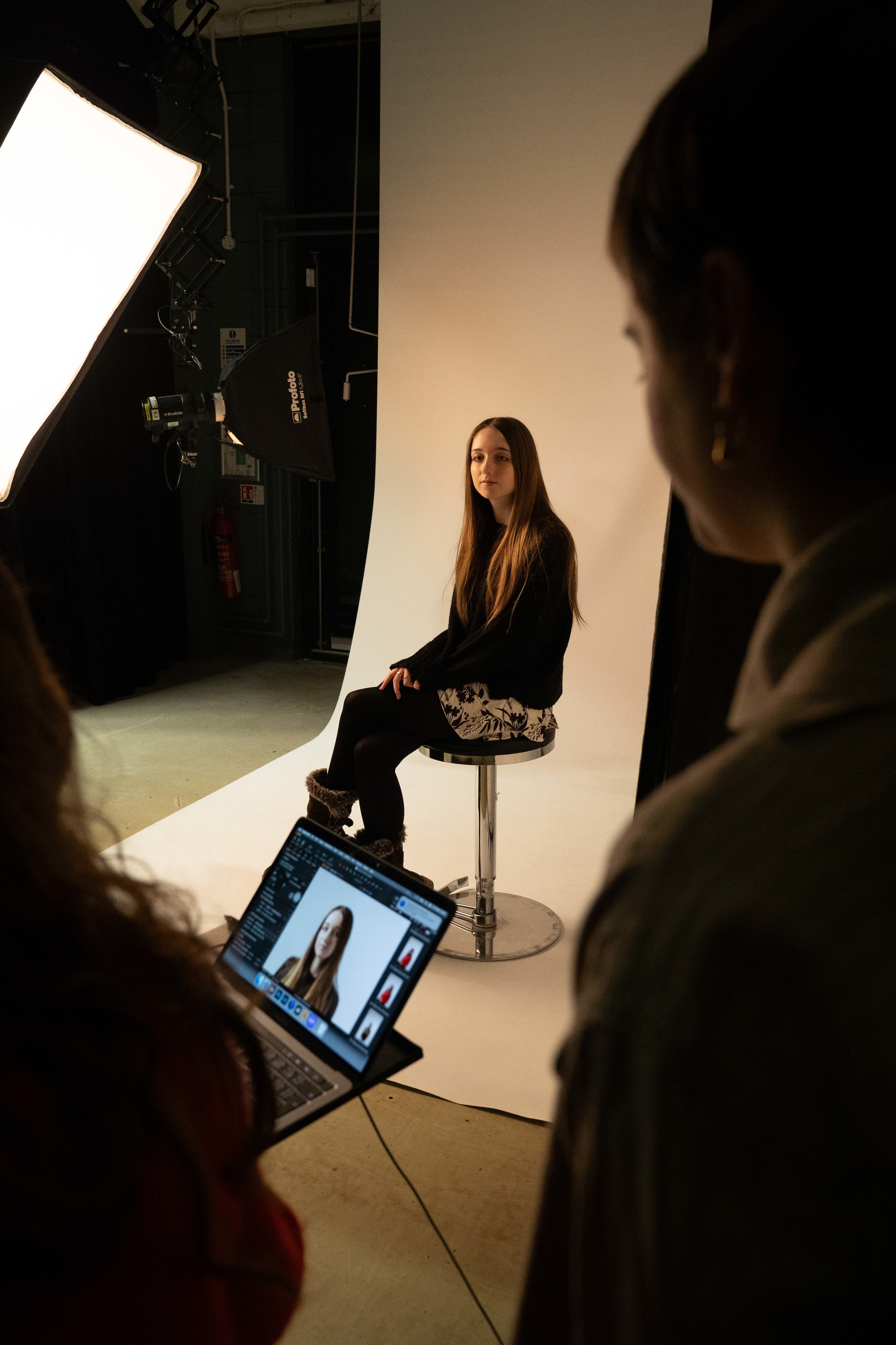 A young person sits on a metal stool and poses for a photo, being taken by other students visible in the foreground. One of them is holding a laptop.