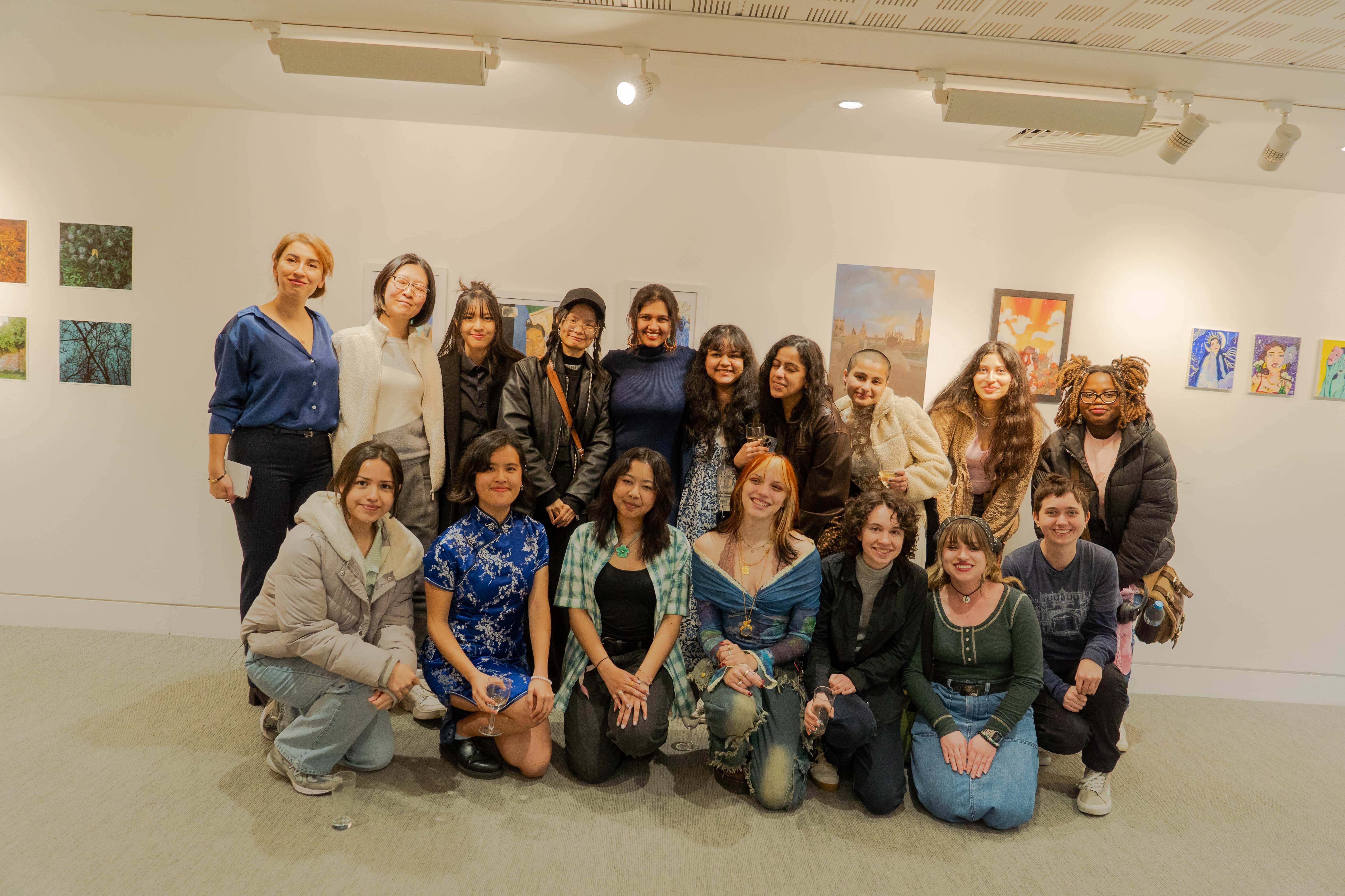 Group of women posing together in white-walled art gallery with colourful paintings displayed on walls and track lighting above.