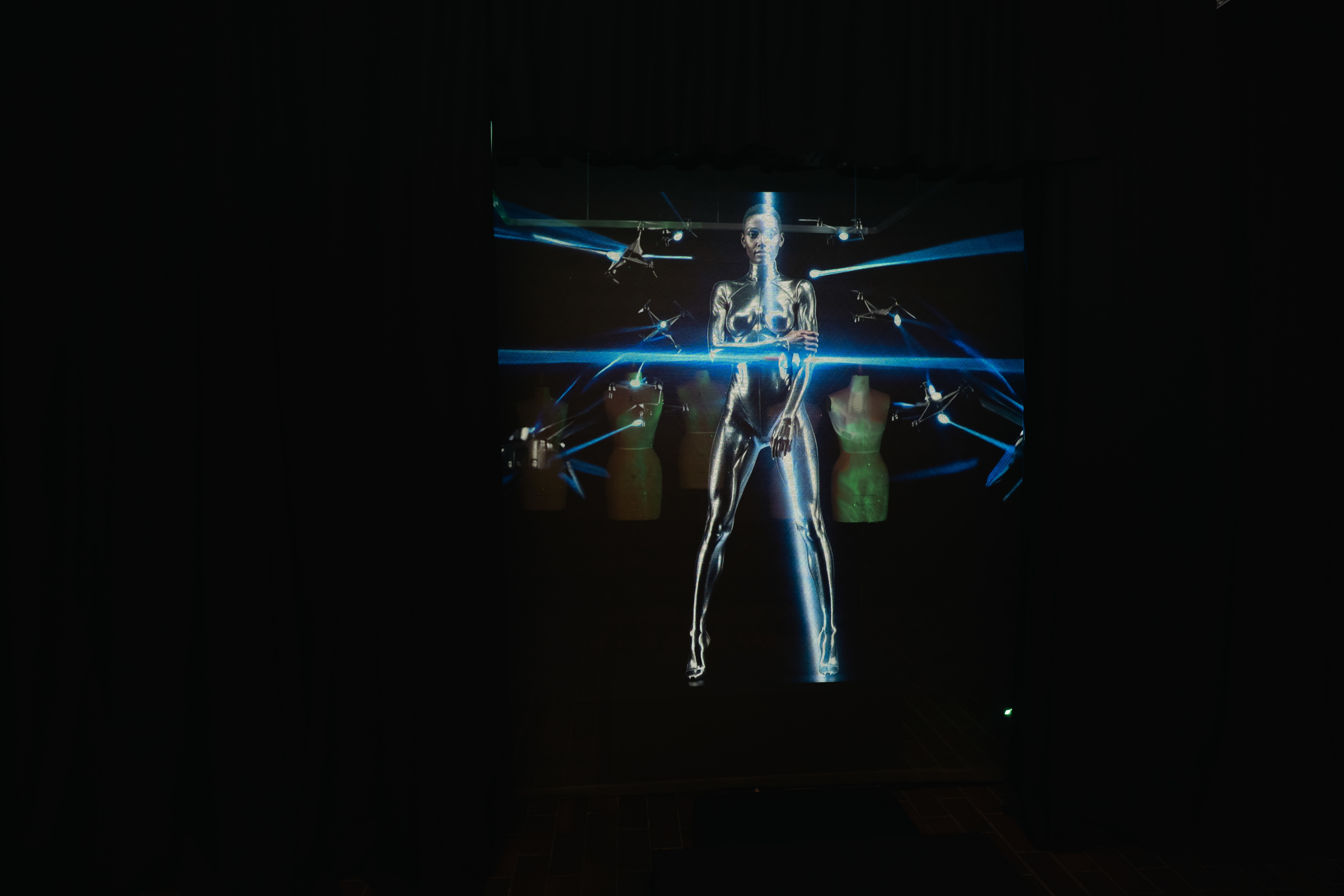 A digital supermodel with spotlights shining in all directions around them, with mannequins visible in the background, appears on a screen in a dark room.