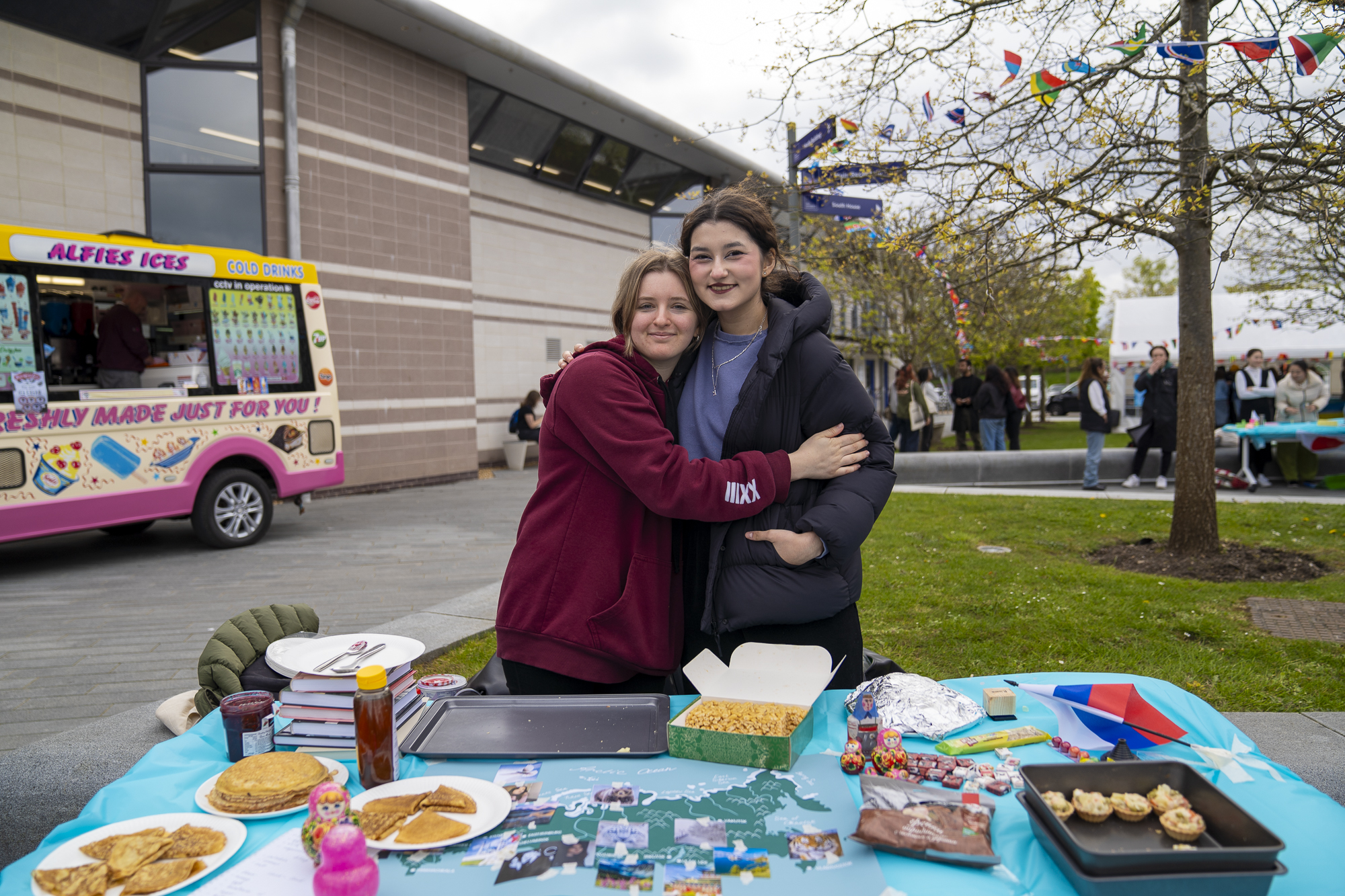 Two students embracing one another in front of a bright blue table with snacks, outdoors on AUB Campus, ice cream van in the background.