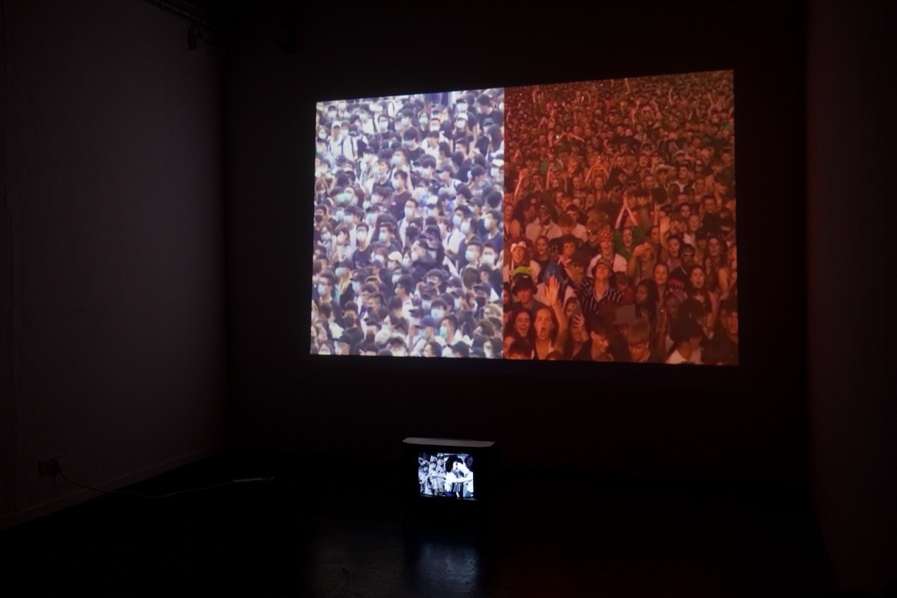 Still shot from RECOMMENDED DOSAGE on a screen with a dark background, crowd of people, the right side has a red filter applied to it. Small screen below displaying more people.