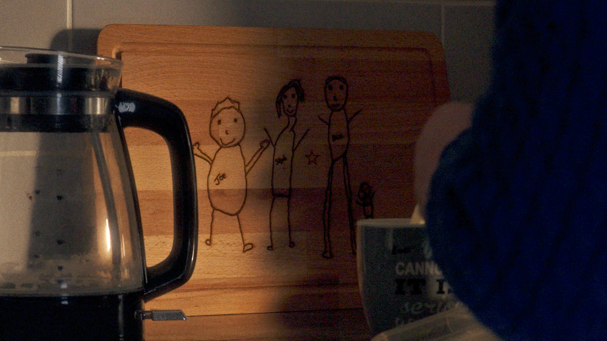 Wooden cutting board with stick figure drawings of people, next to chrome kettle and mug in dimly lit kitchen.