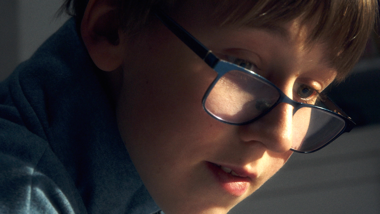 Person wearing black-framed glasses looking downwards, warm lighting illuminating face from below, wearing dark blue clothing.