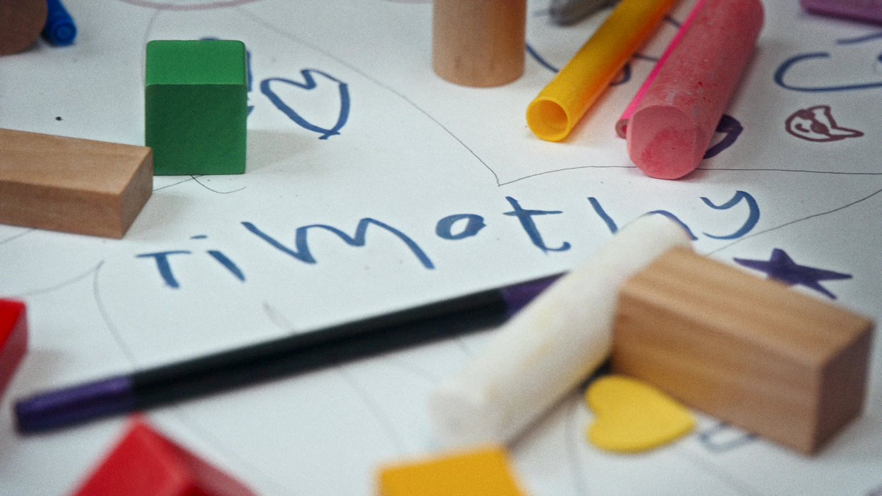 Children's educational materials on white paper: wooden blocks, coloured pencils, and handwritten letters including "Timothy".