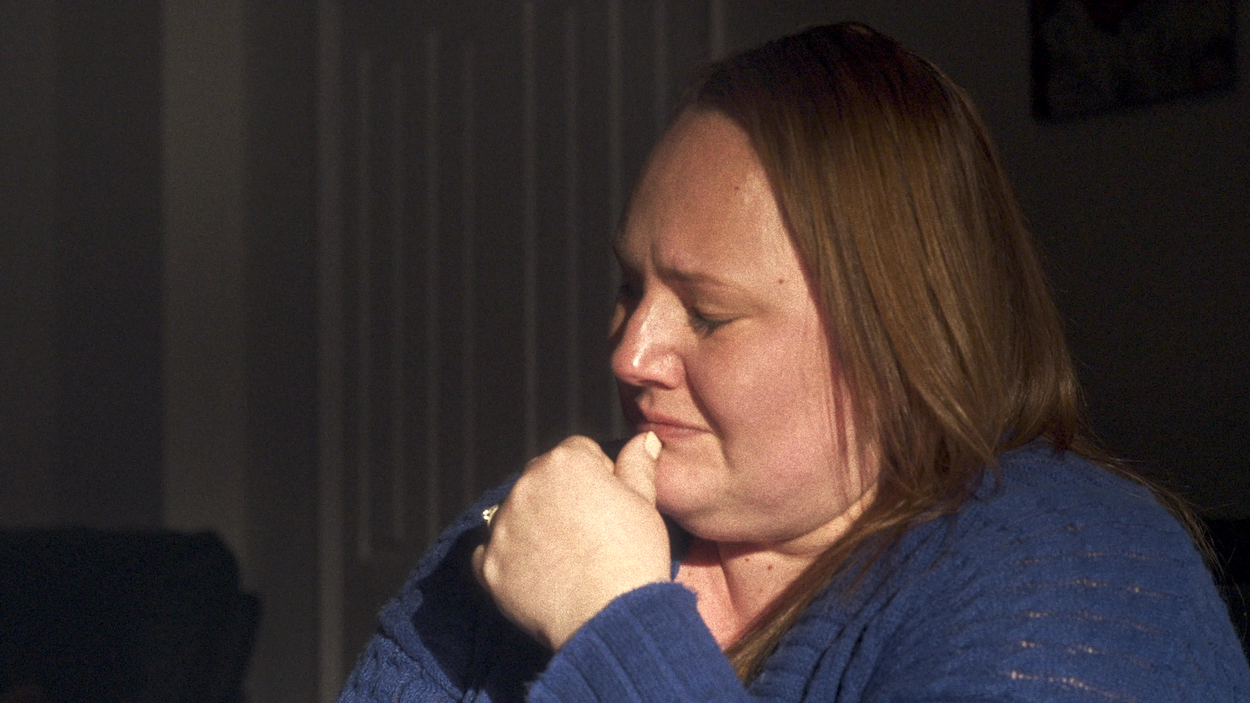 Woman with shoulder-length brown hair in blue jumper, hand positioned near chin, photographed in profile against dark background.