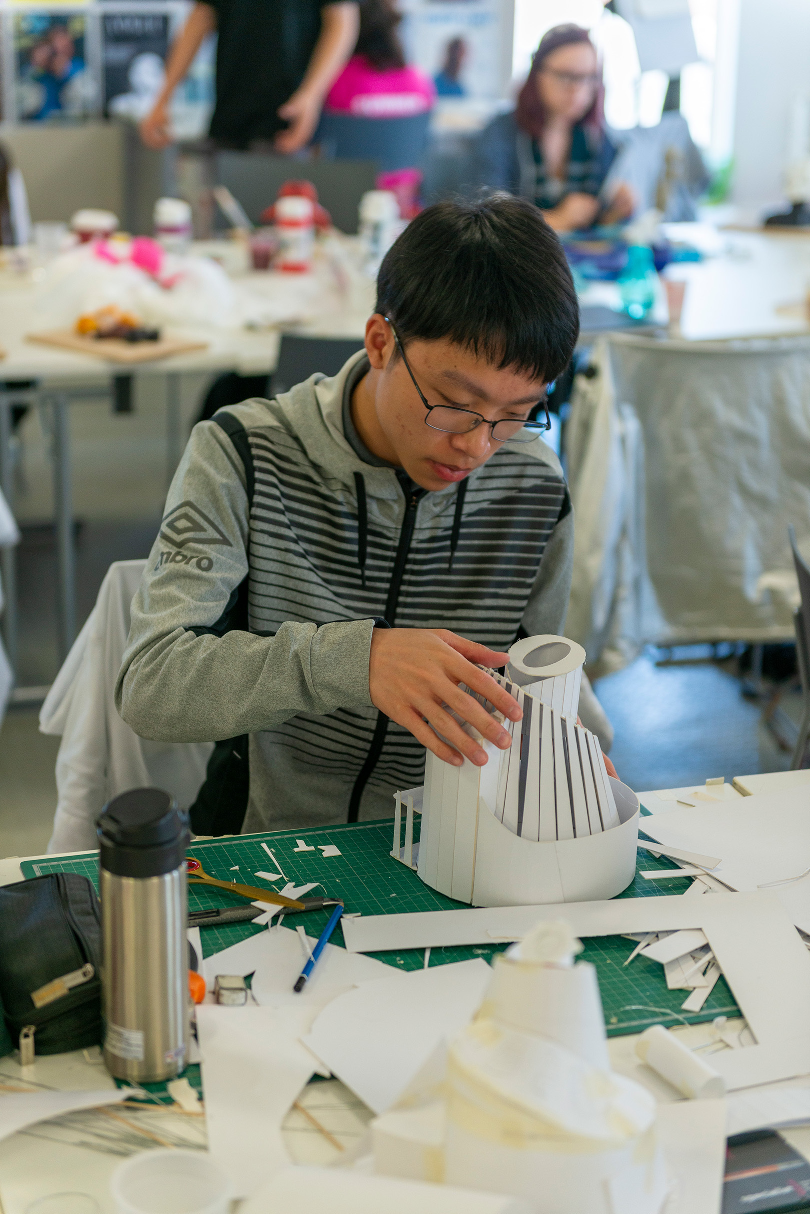 Student sitting at a table with a cutting board in front of them and working on an architectural model made of card.