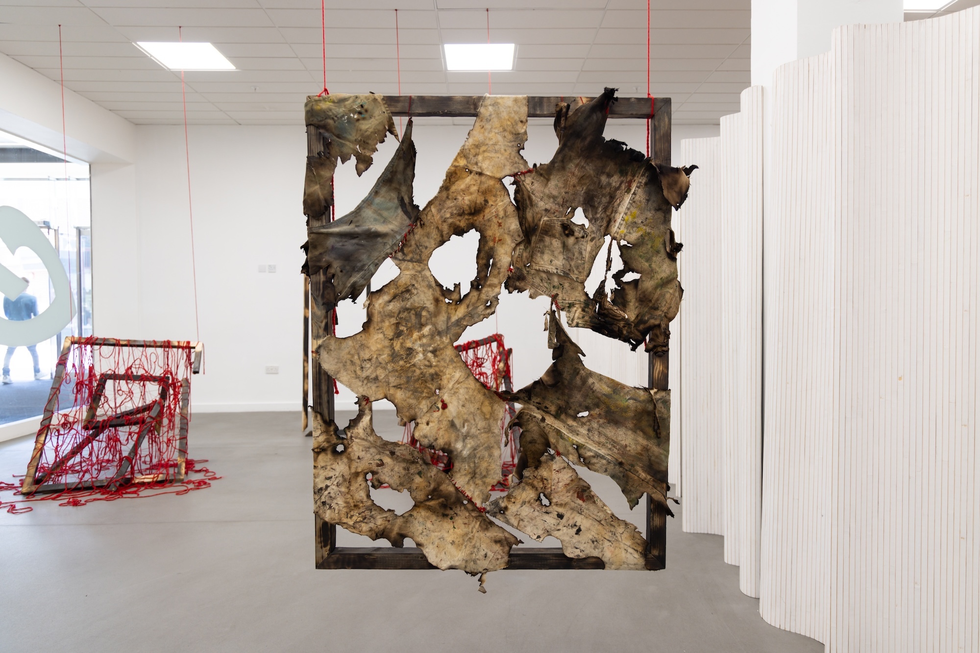 Gallery installation with suspended rectangular frame containing brown camouflage fabric with white angular shapes attached. Red sculptural elements visible in background.