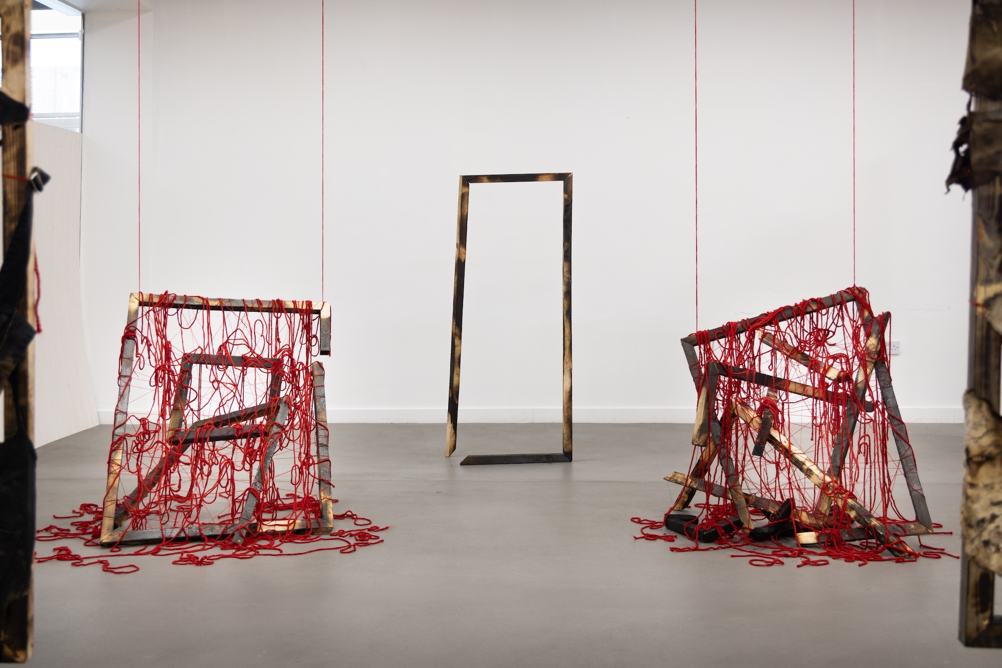 Gallery installation with two red wire sculptural structures flanking a tall rectangular mirror frame against white panelled wall.