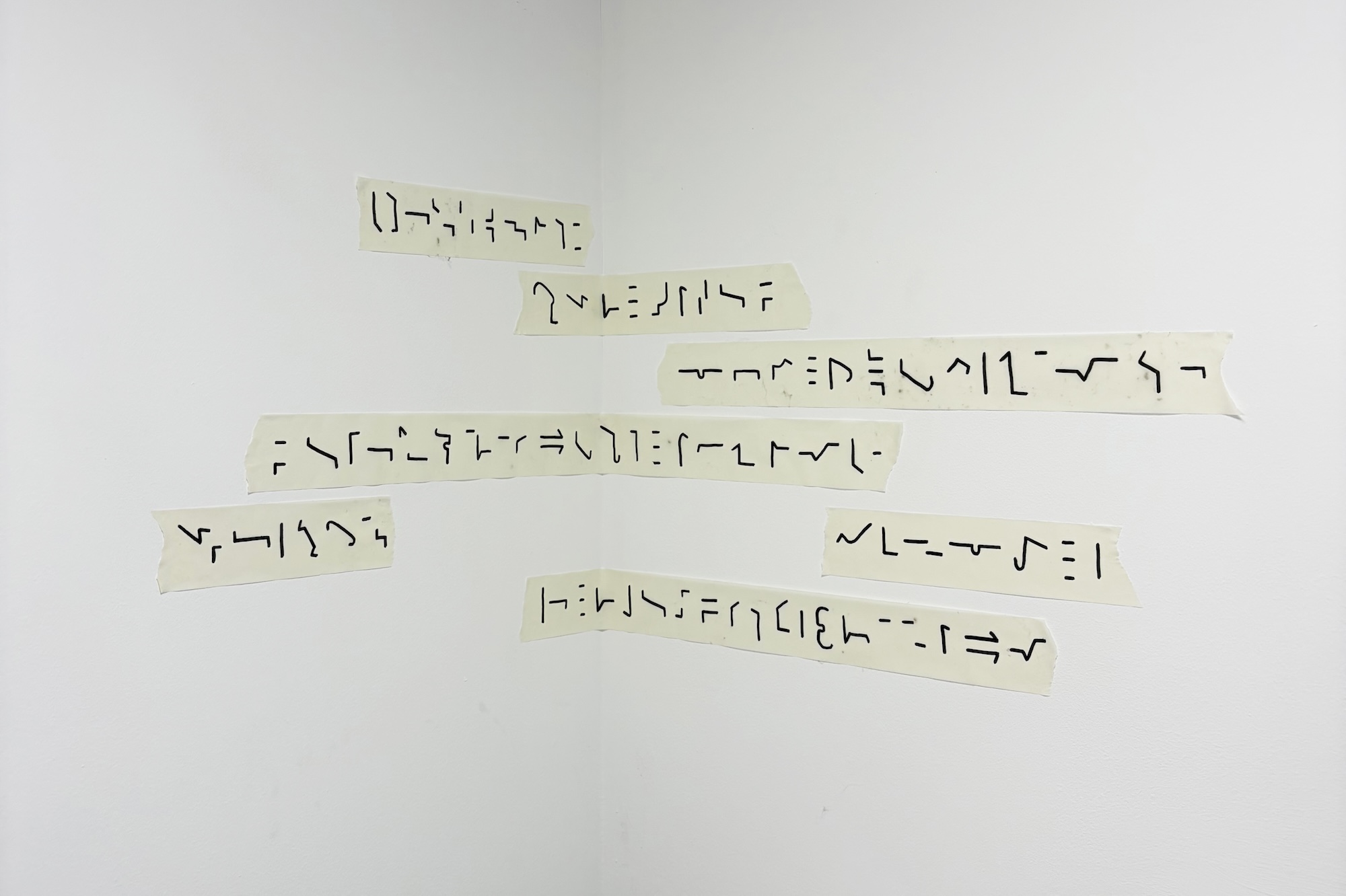 Several strips of paper with abstract symbols are pasted on a white wall, arranged in a non-linear pattern. The symbols vary in design and size, with some resembling lines and curves, creating an unconventional visual display.