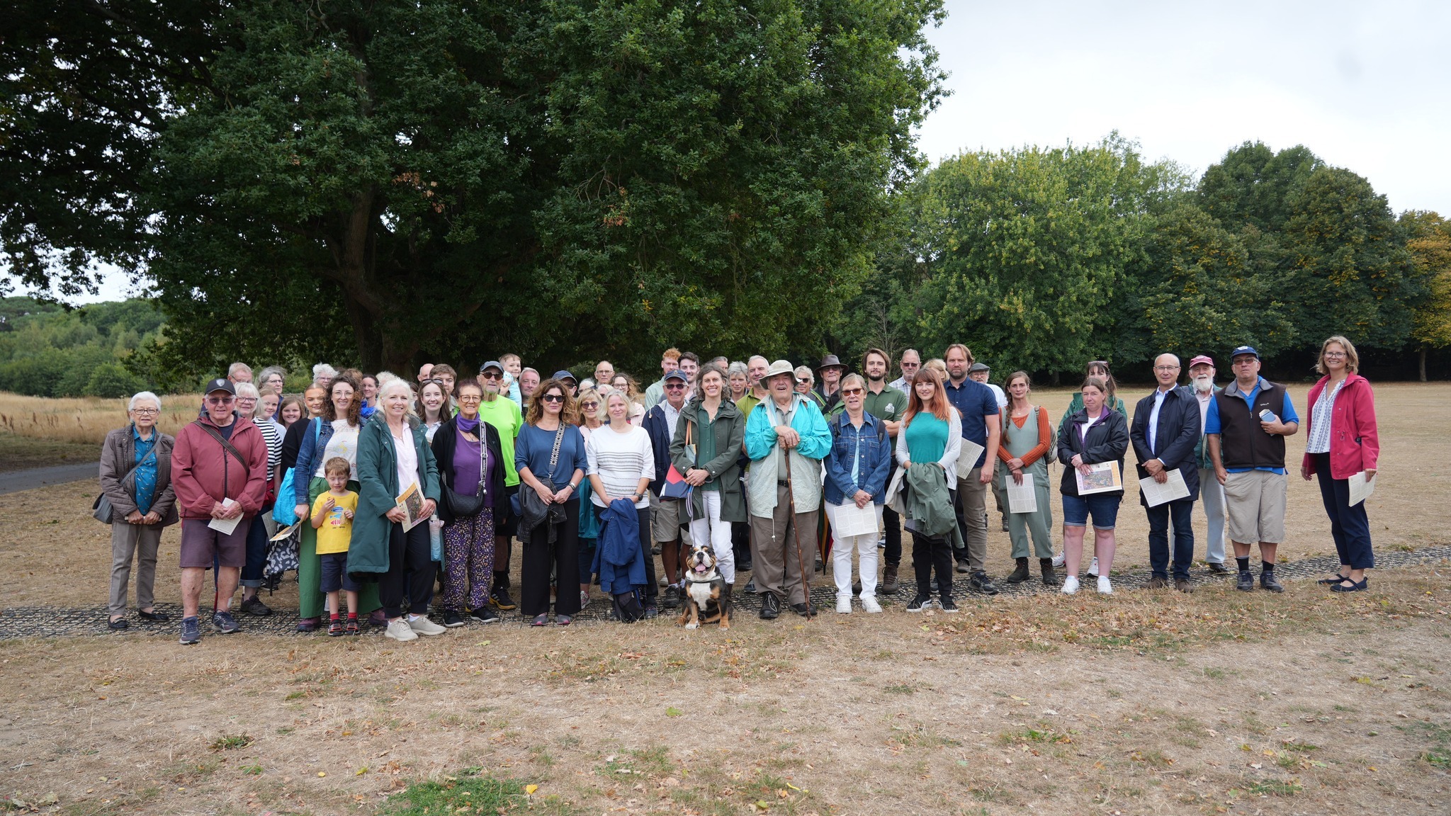Large group of people standing together on bare ground with dense green trees behind them and cloudy sky above.