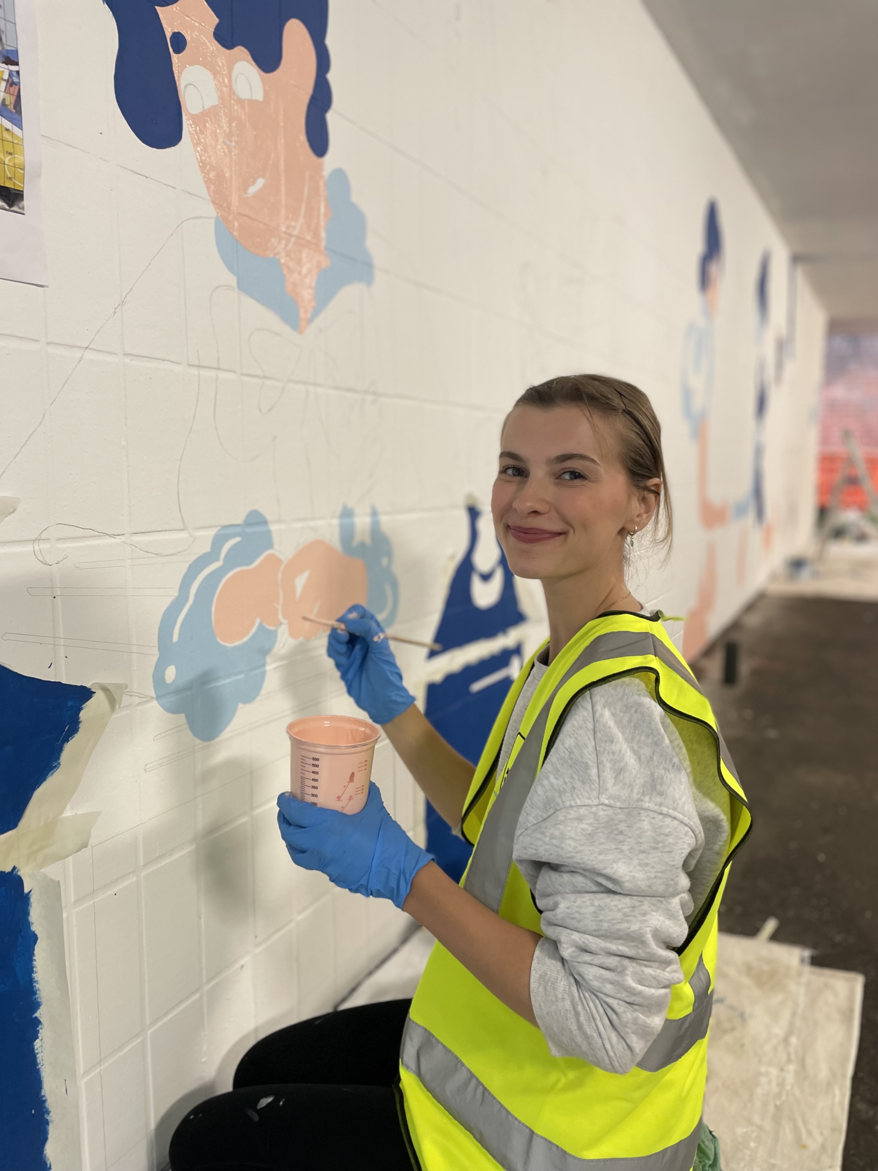 A BA (Hons) Graphic Design student in a yellow hi-vis vest and blue gloves painting colourful mural with peach, blue and navy shapes on white wall.