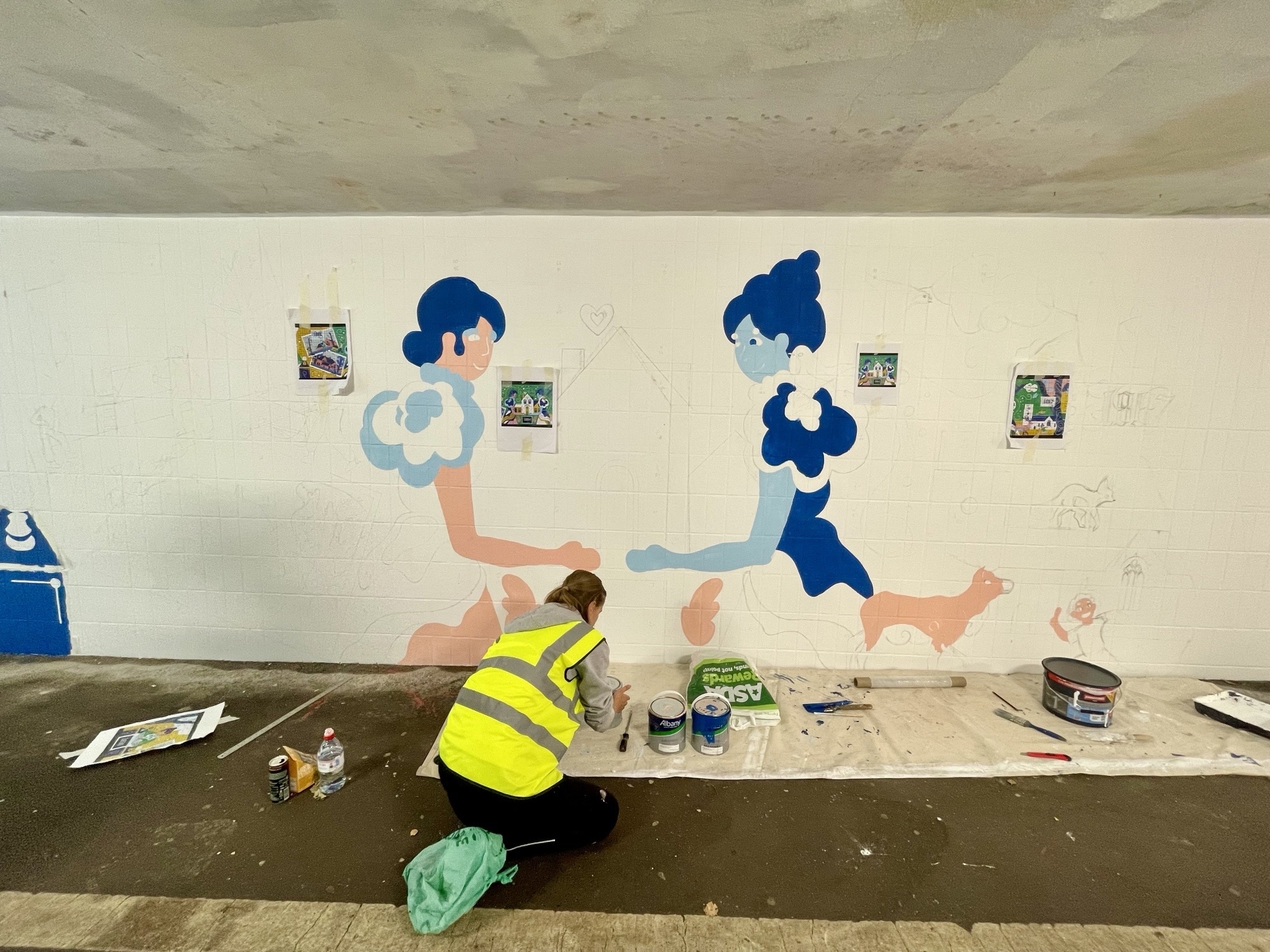 A BA (Hons) Graphic Design student in hi-vis jacket painting mural on white wall featuring two blue-haired figures. Paint pots and materials scattered on floor below.
