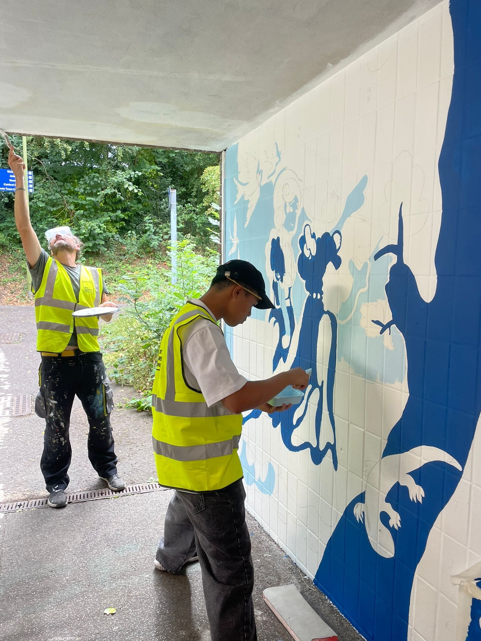 Two BA (Hons) Graphic Design students in hi-vis yellow vests painting a mural with blue figures on white wall under covered area near trees.