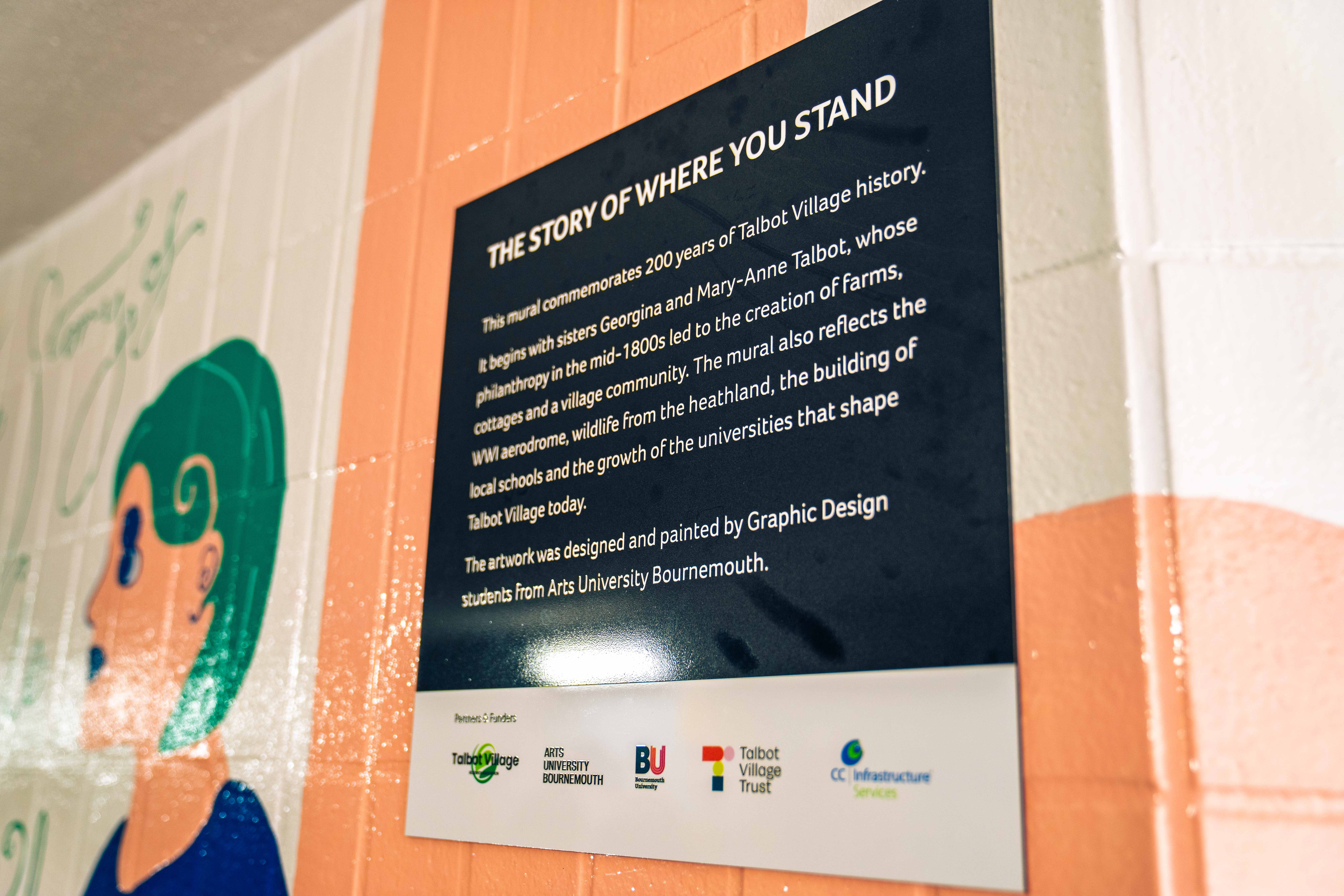 Black museum information plaque titled "THE STORY OF WHERE YOU STAND" mounted on orange and white painted wall with partial mural visible.