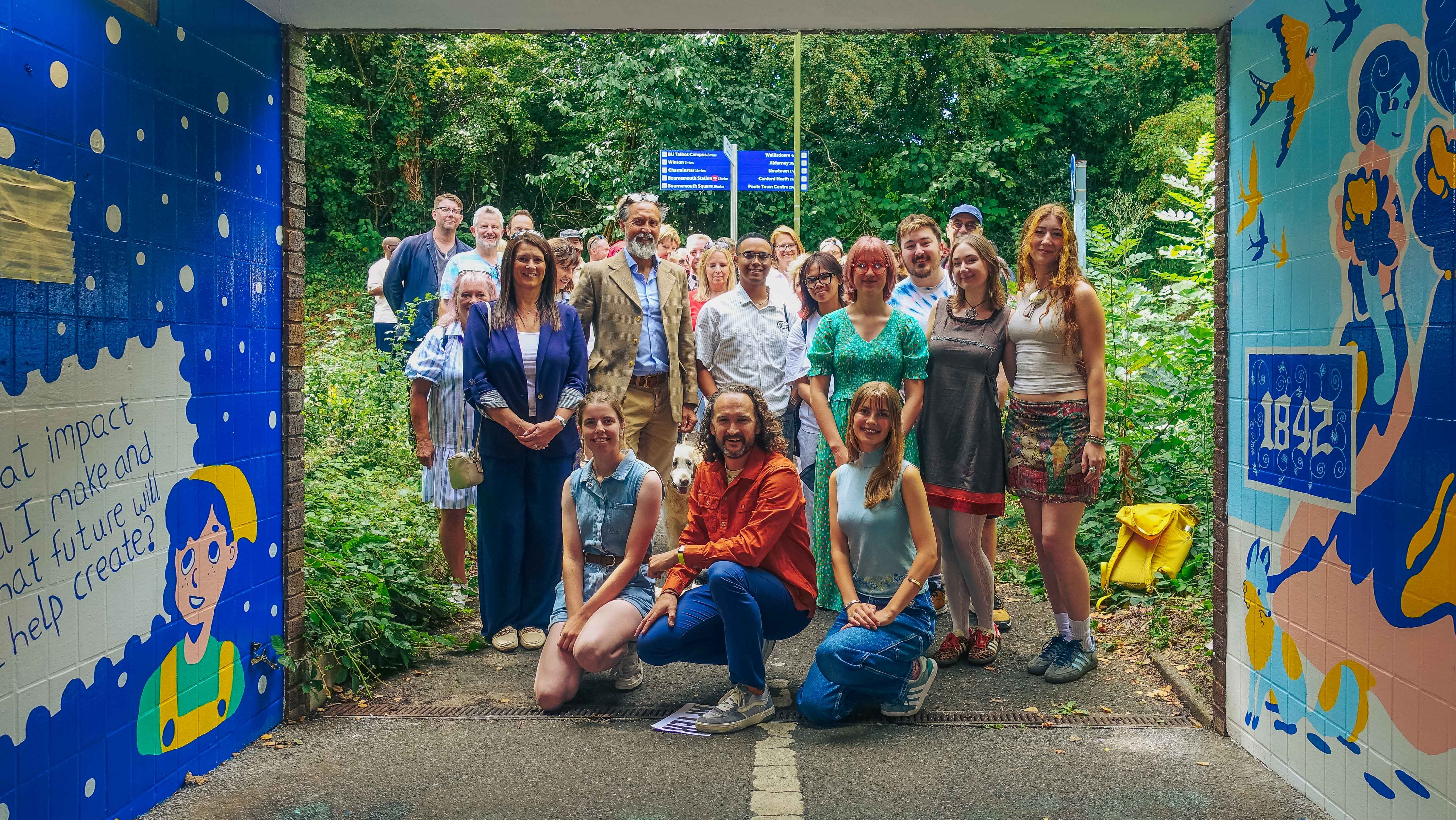Group of people including staff and students of Arts University Bournemouth posing at colourful blue-painted underpass with mural artwork, surrounded by green trees and foliage.