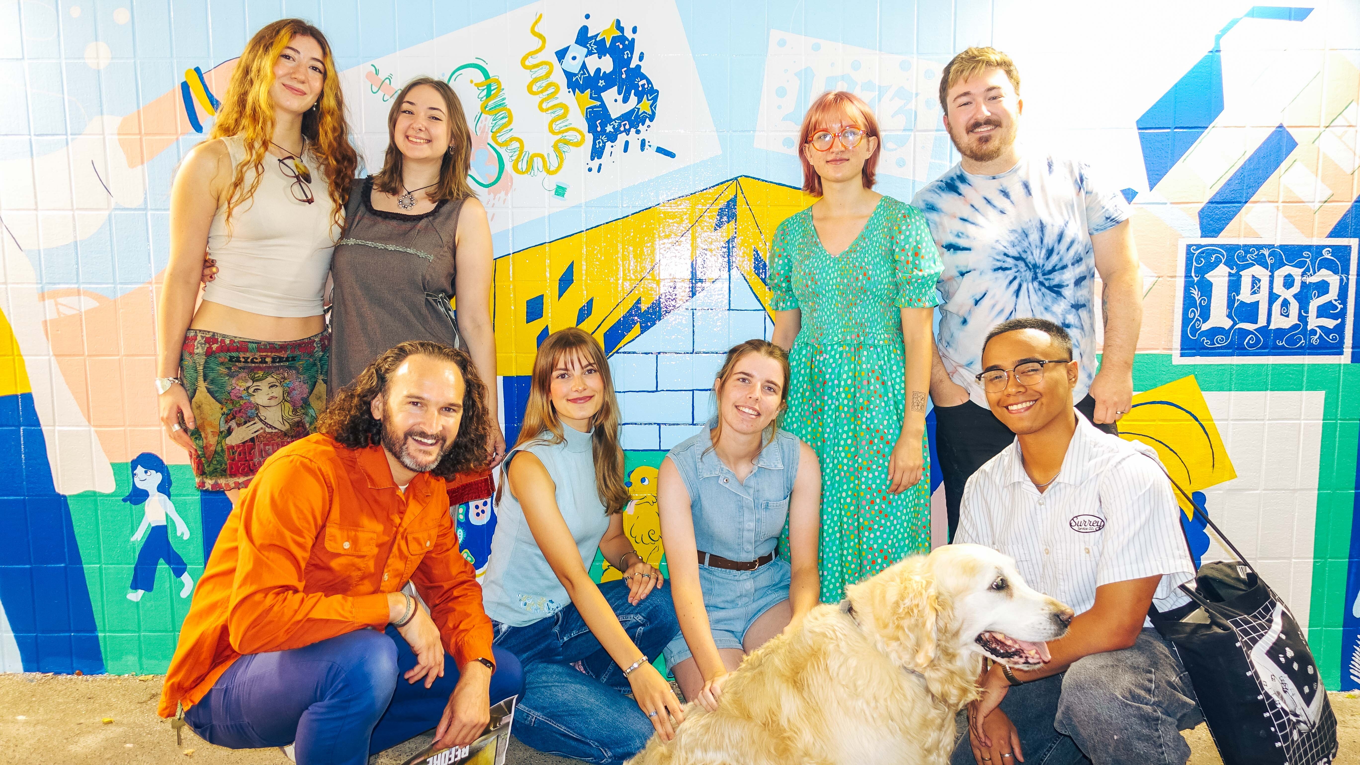 BA (Hons) Graphic Design students and M Sims posing with golden retriever in front of colourful mural featuring blue, yellow, and white geometric shapes and "1982" text.