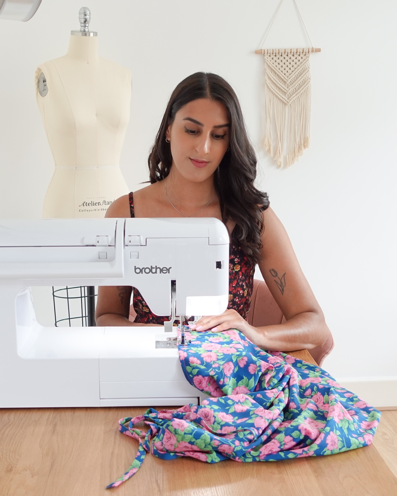 T Johal using white Brother sewing machine on colourful floral fabric. Dress form and macramé wall hanging visible in background.