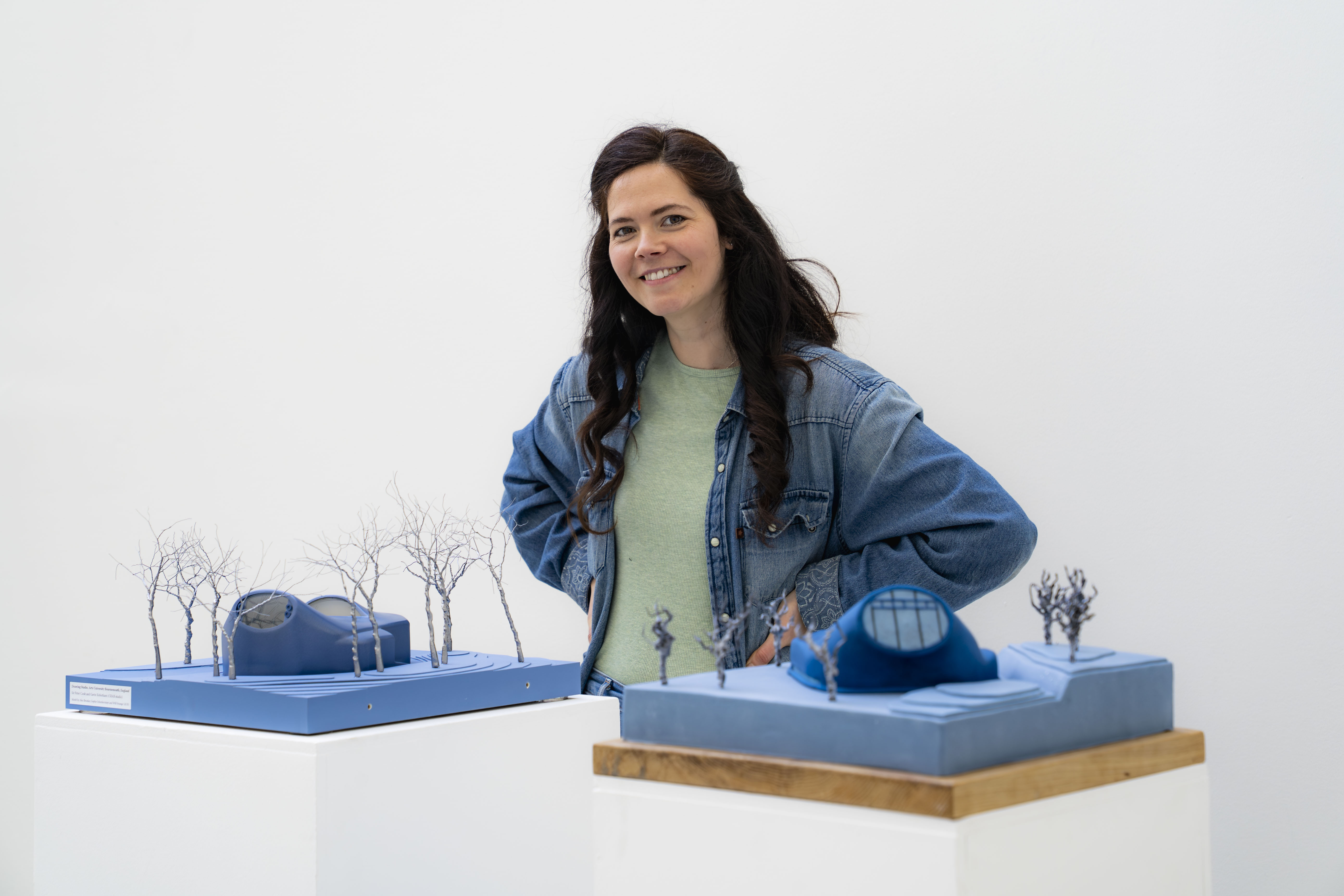 Woman in denim jacket and mint top stands behind two blue architectural models with miniature trees and buildings on white surface.