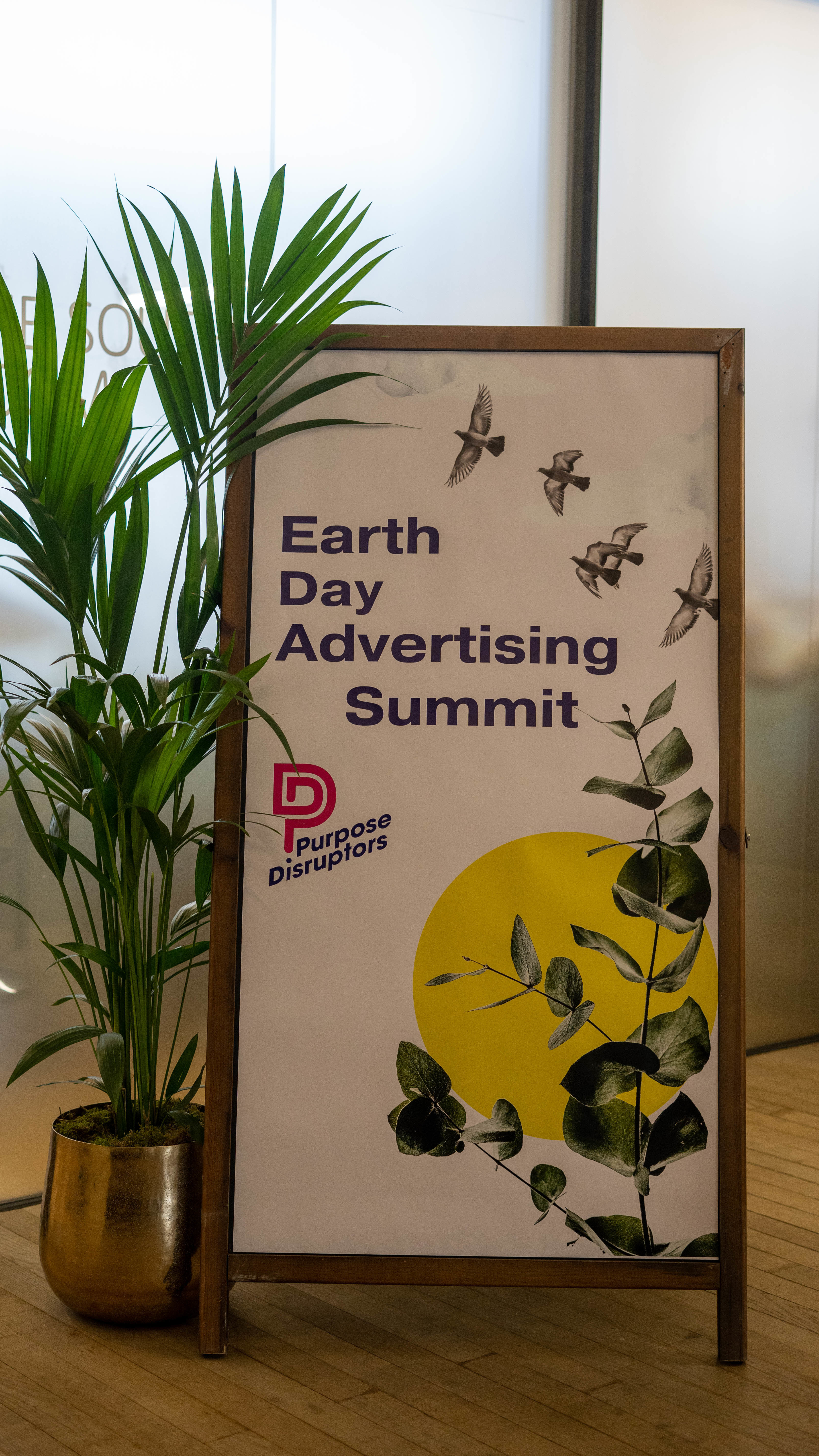 A-frame with a poster with the text "Earth Day Advertising Summit" is displayed next to a plant.