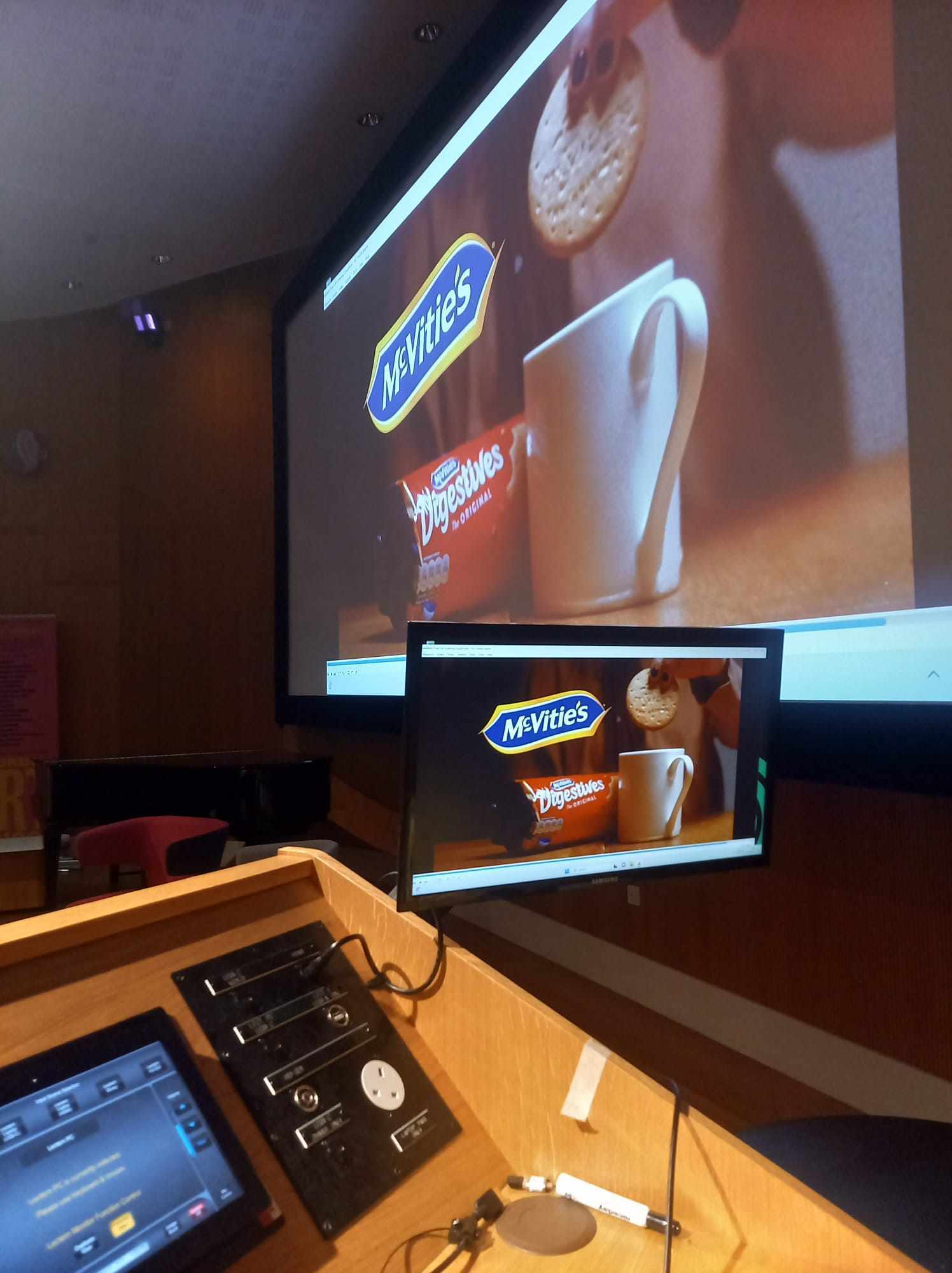 A lecture theatre lectern showing a screenshot of the AUB BA (Hons) Film Production McVitie's commercial 'Tea Rebellion' on the monitor, and on the big screen in the background.
