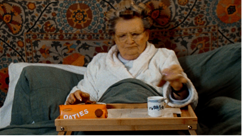 Still from BA (Hons) Film Production student commercial "Tea Rebellion", showing an older woman in a dressing gown sitting on a sofa with a tray table over her lap, about to dunk a biscuit into a cup of tea.
