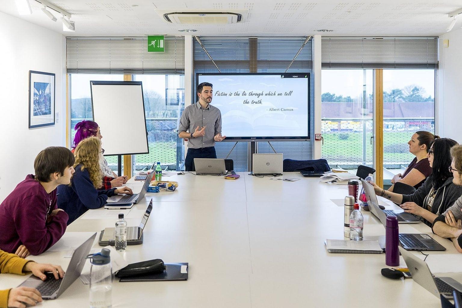 A person stands at the head of a table in front of a screen and delivers a lecture to people sitting either side.