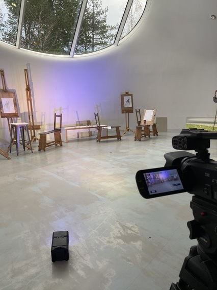 A collection of wooden furniture and artists' easels in the AUB Drawing Studio. A camera appears in the foreground.