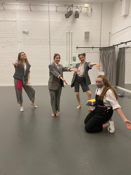 Three BA (Hons) Dance students posing in a studio. They are holding out their arms presenting a fourth person, kneeling on the right-hand side.