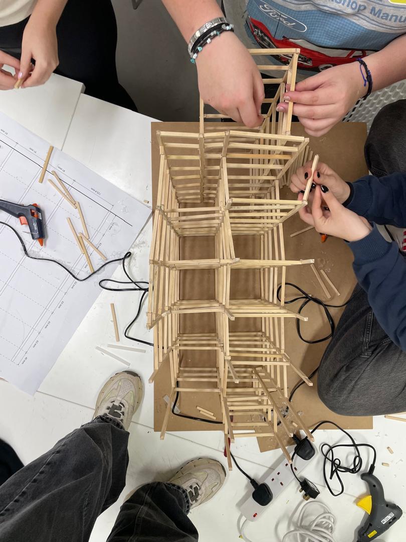 Students building wooden tower structure with multiple levels, working collaboratively with plans and tools on table.