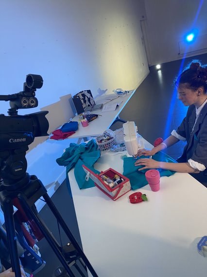 A person sitting at a table using a sewing machine. A camera is set up on the left-hand side pointed down at them.