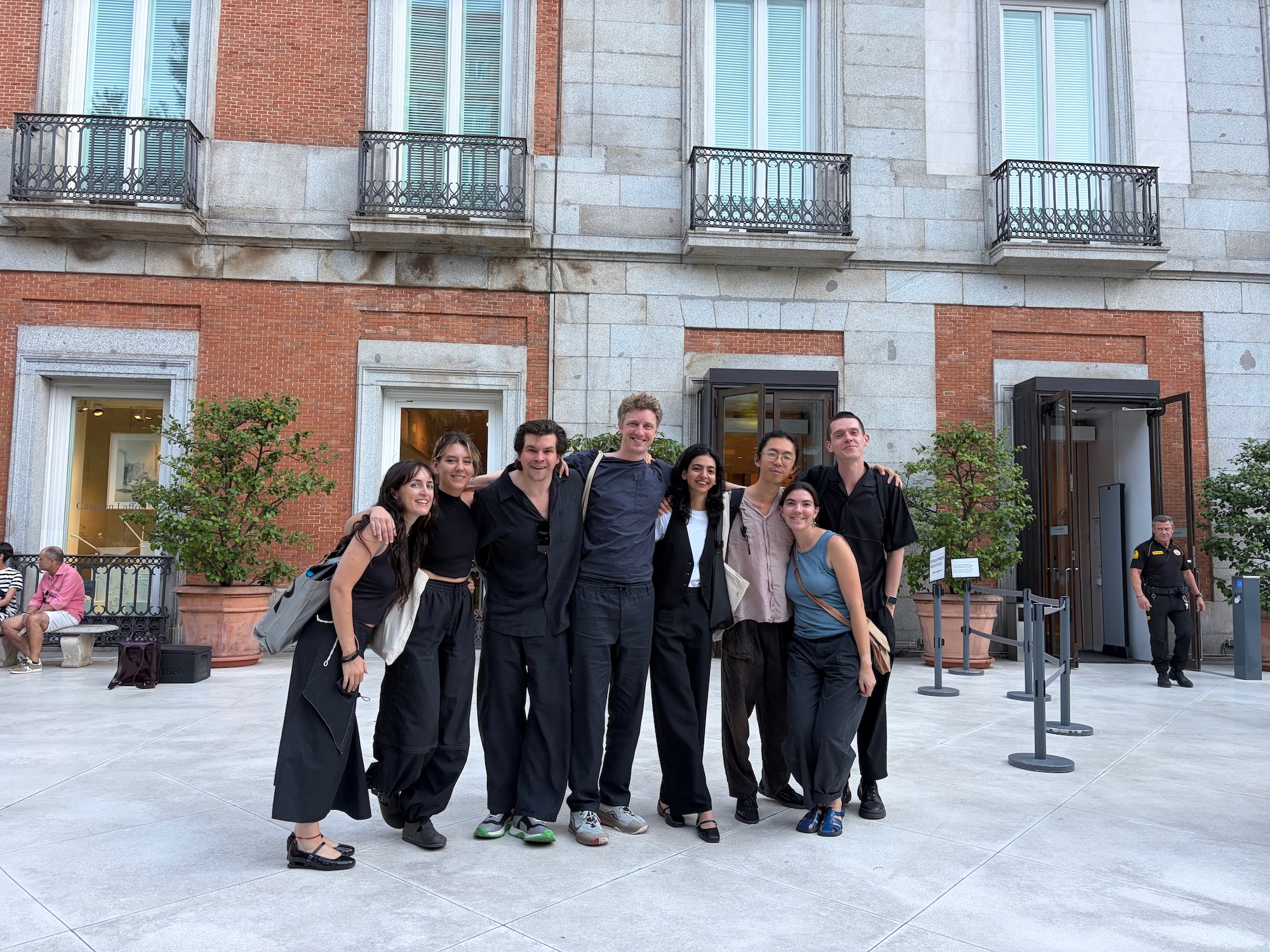 A group of nine individuals posing together outside a building with large windows and balconies. They are dressed in black attire, with some wearing lighter shades. The background features brick walls and potted plants.