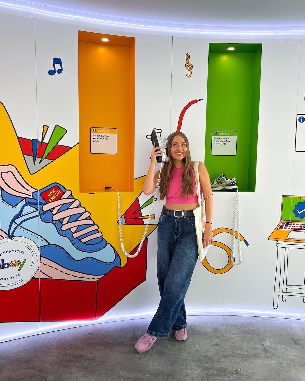 Tegan Price stood with a shoe up to her ear meant to be a phone, stood in front of her own colourful illustrations of sneakers in ebay logo colours.