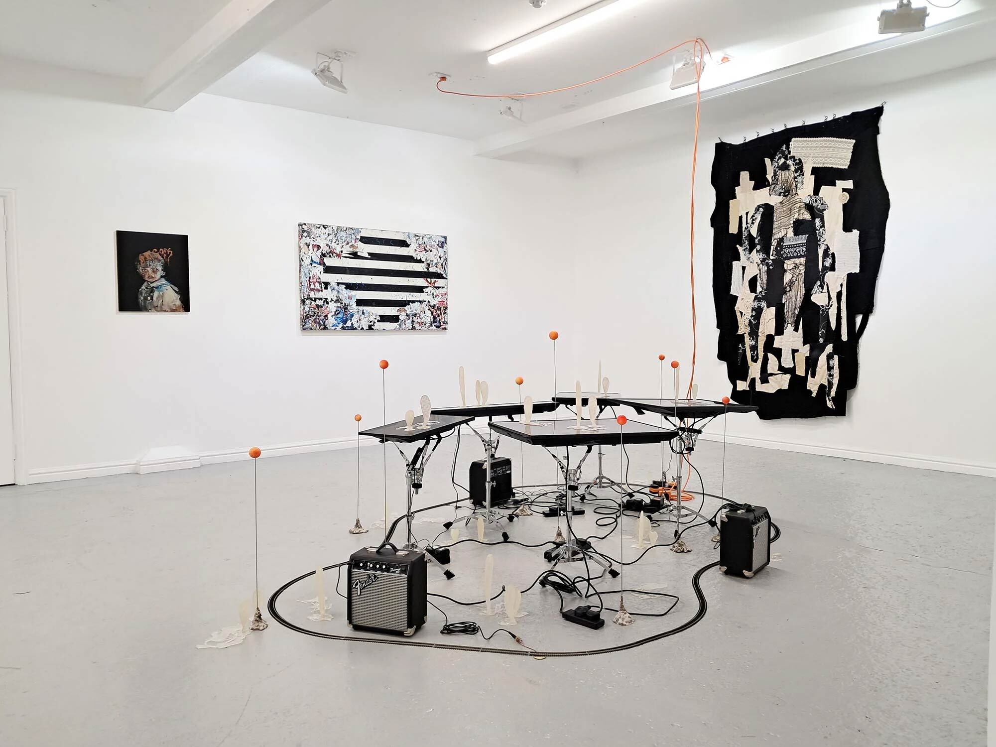 White gallery space with electronic installation featuring cables, amplifiers and metal table in centre, three artworks on walls including black abstract piece.
