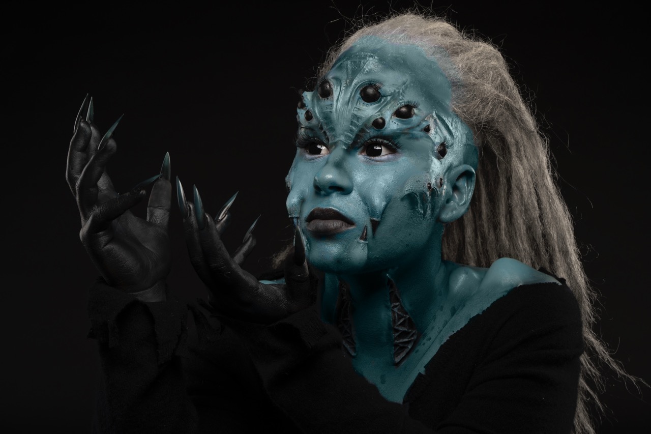 Kasandra Georgi in alien-like sea creature make-up, turquoise with blonde hair and black hands.