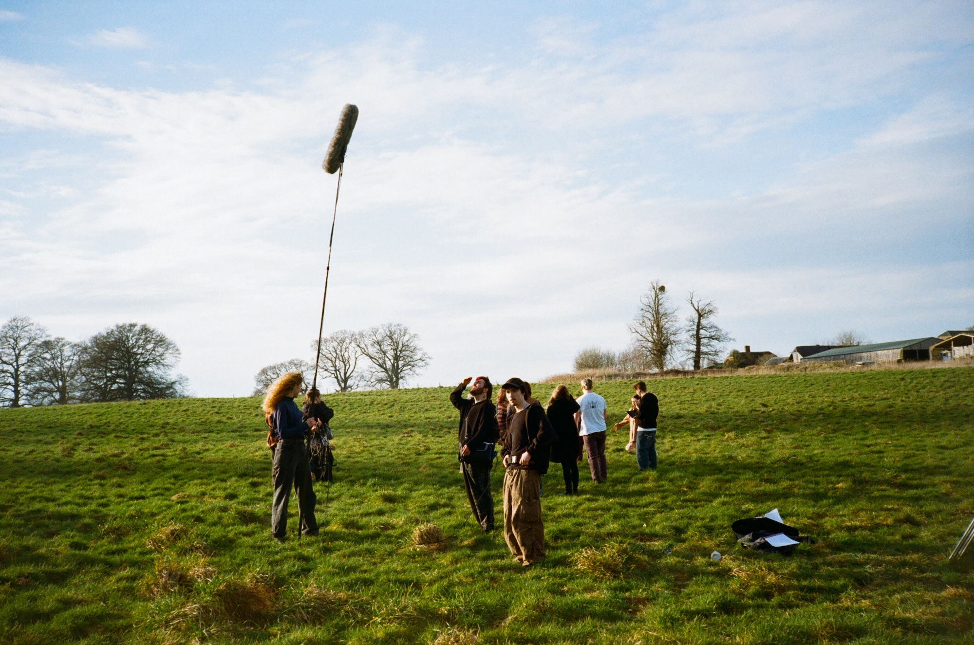 BA (Hons) Film Production students crew with boom microphone and equipment filming in green field under cloudy sky, bare trees and buildings in background.