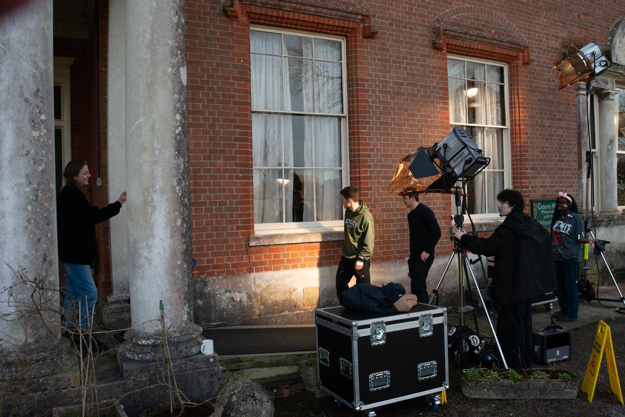 Crew of BA (Hons) Film Production students setting up equipment outside red brick building with white columns, lighting gear and cameras positioned on courtyard.
